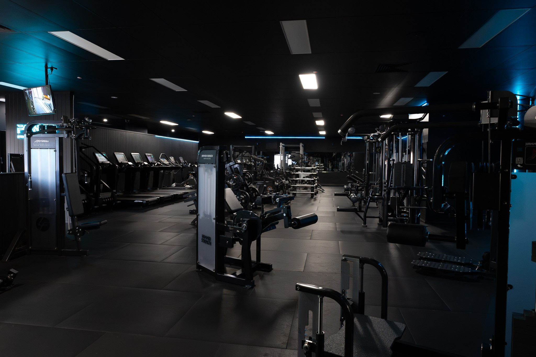 Interior of a modern, dimly lit gym with various workout equipment, including treadmills and strength training machines, illuminated by overhead lights.