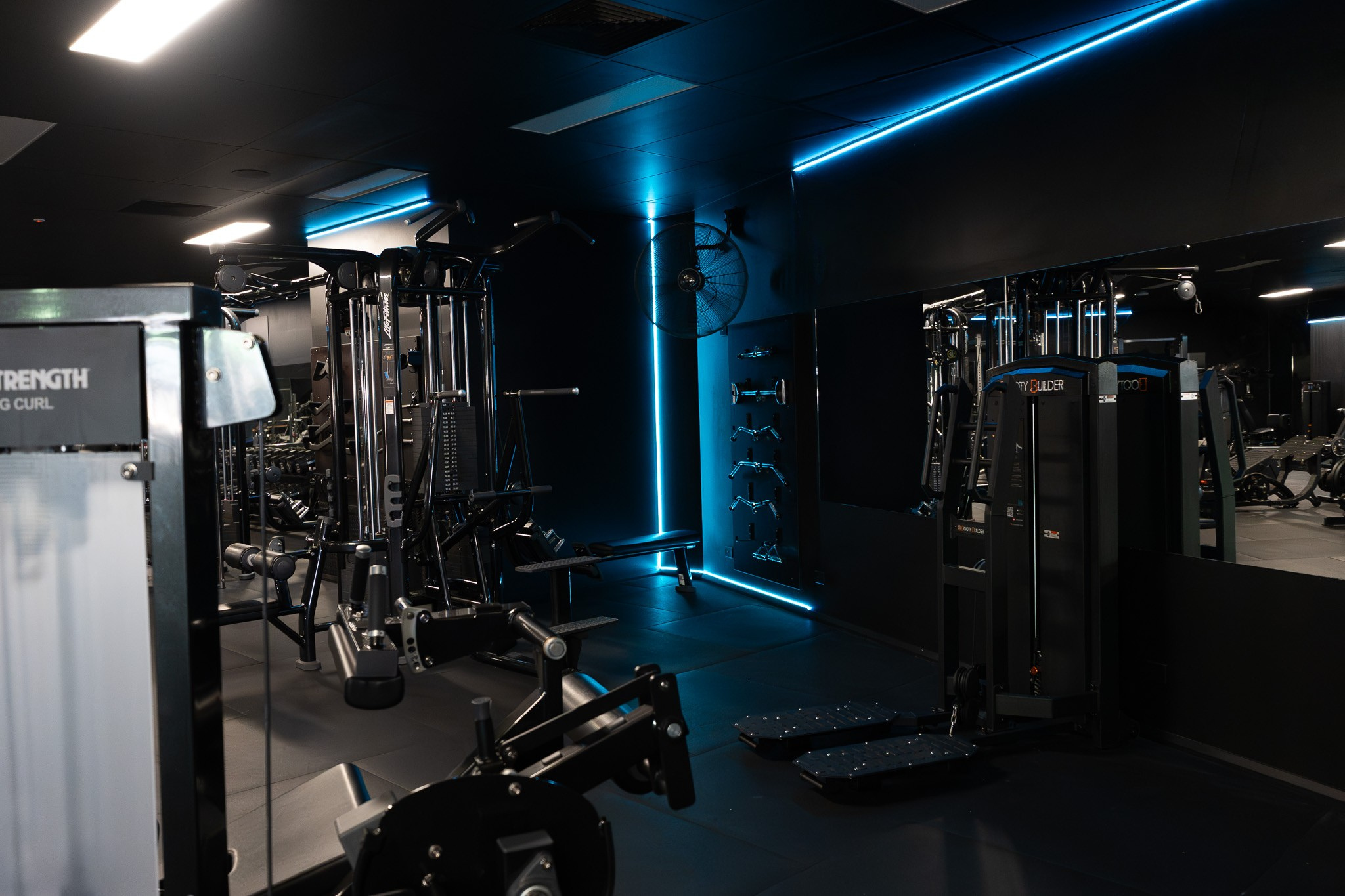 A modern gym with exercise equipment, black walls, blue LED lighting, and a wall-mounted fan.