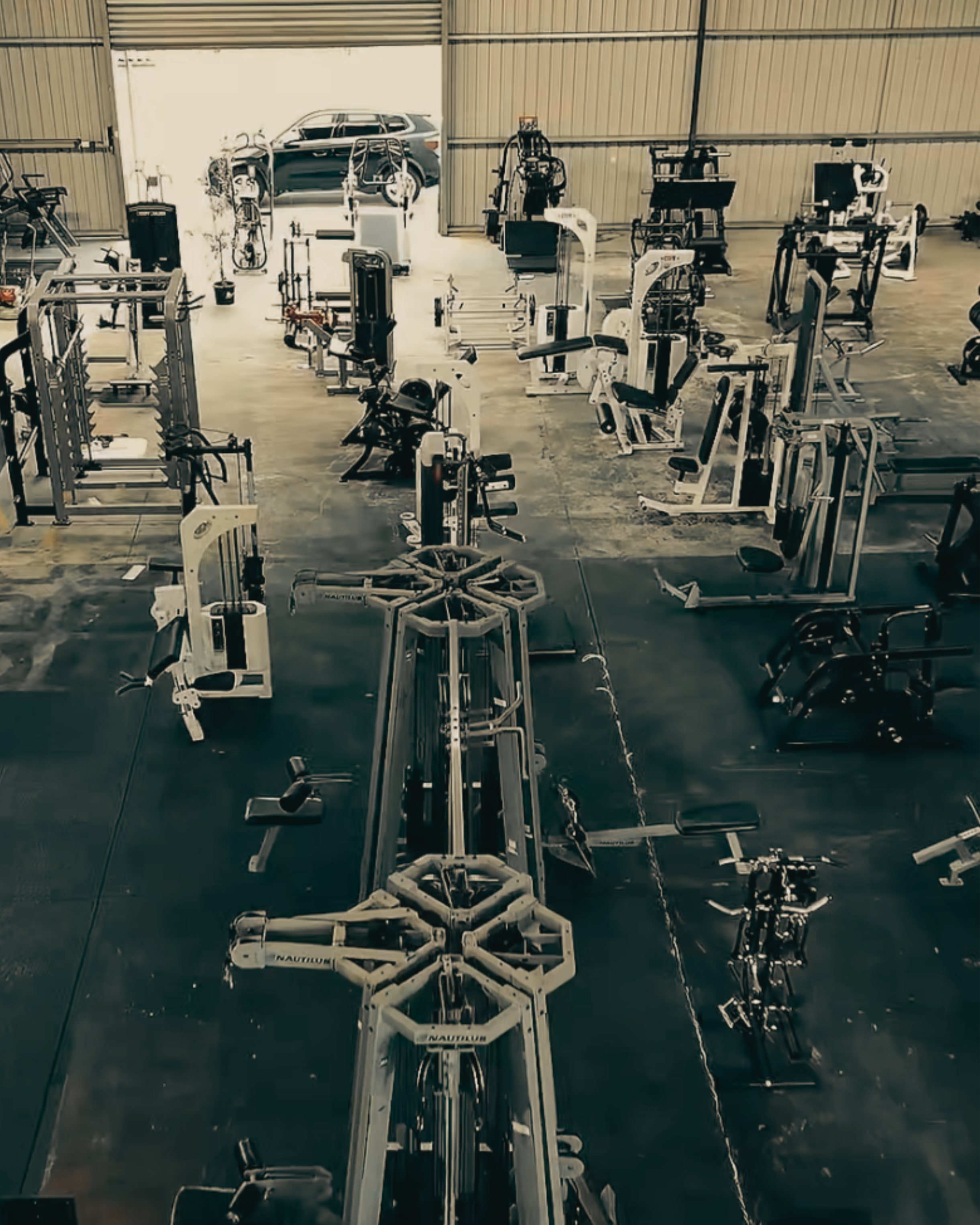 Indoor gym with various exercise equipment including rowing machines, a weight machine, and a bicycle, with an open garage-style door revealing a parked vehicle outside.
