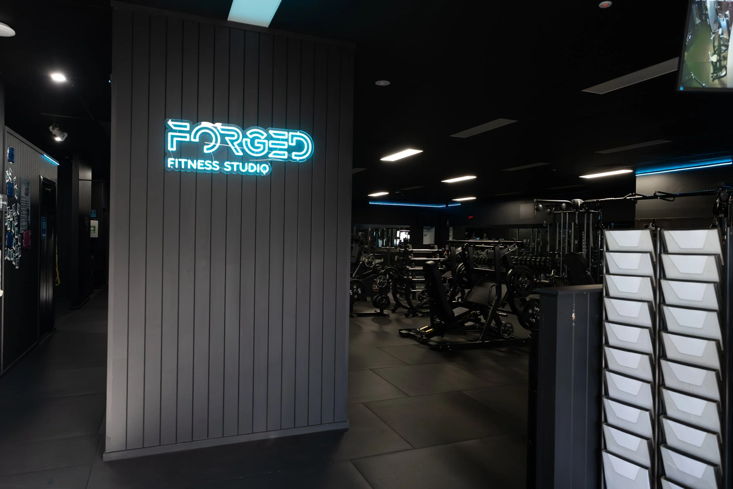 Interior of a fitness studio with a neon sign reading 'FORGED FITNESS STUDIO,' gym equipment including weights and machines, and dark color scheme with modern lighting.