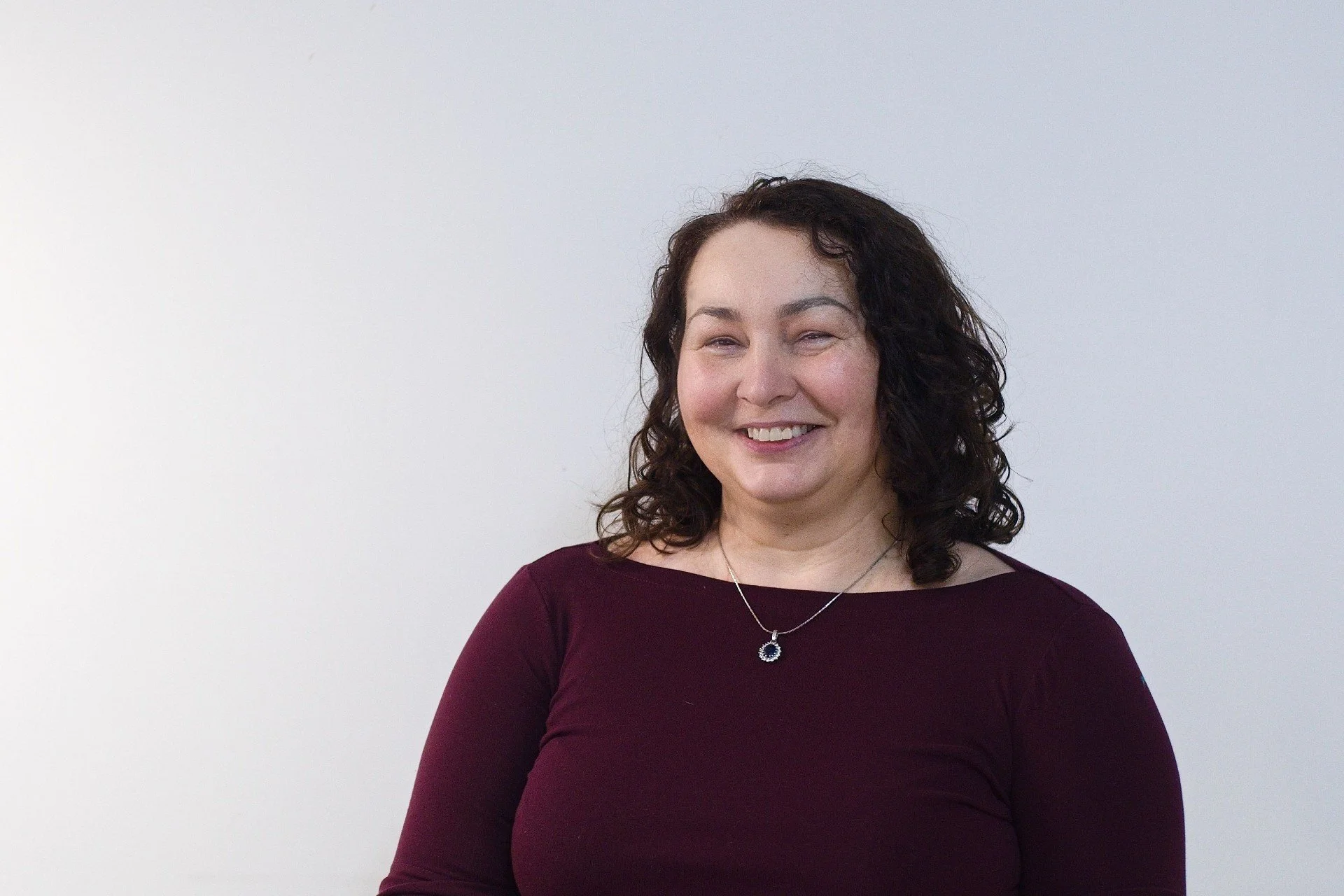 A woman with curly dark hair wearing a dark purple top and a necklace, standing against a plain white background, smiling.