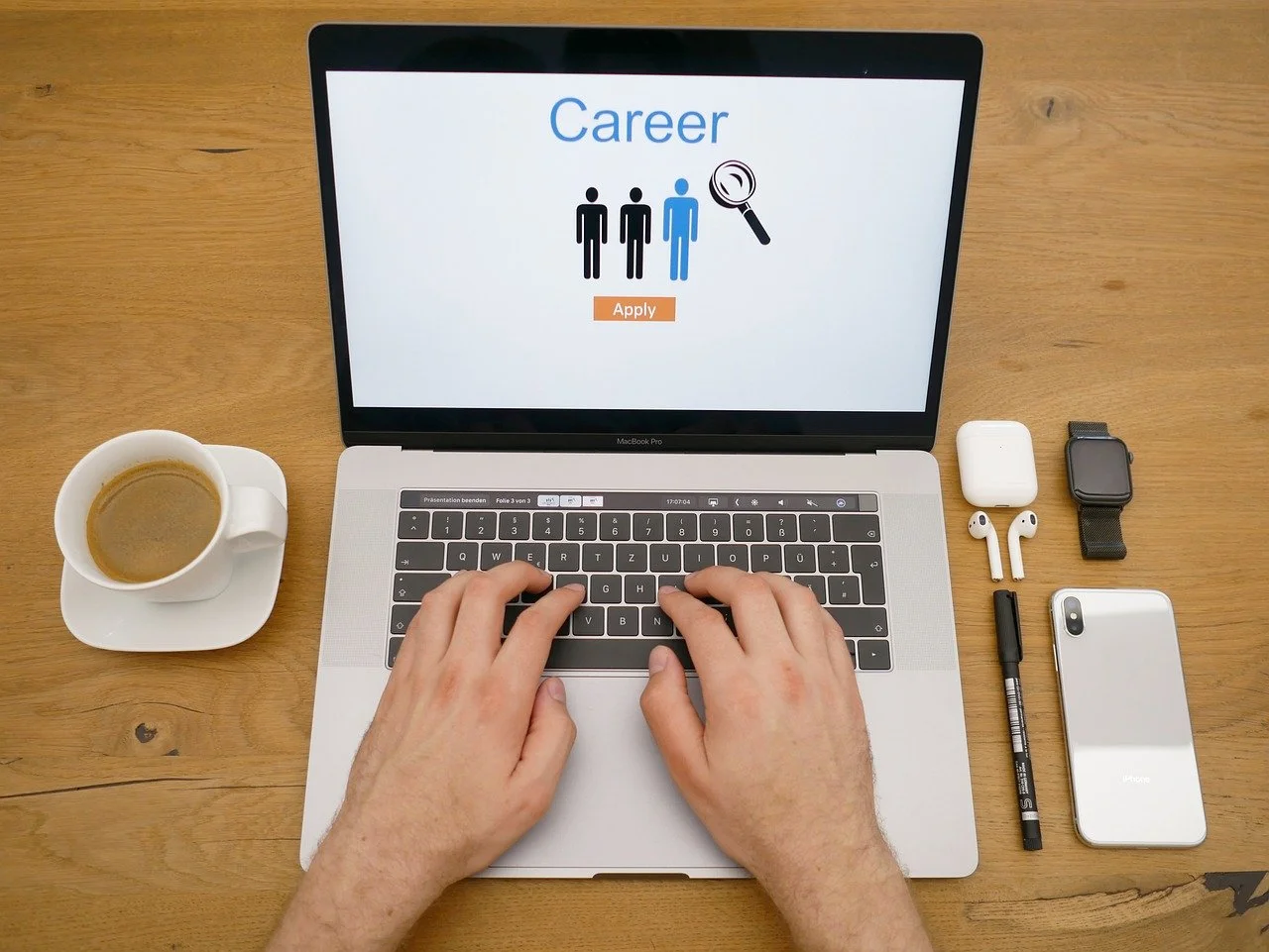 A person typing on a MacBook Pro at a wooden desk with a coffee cup, AirPods, AirPods case, smartwatch, black marker, and white iPhone nearby. The laptop screen displays a career application webpage.