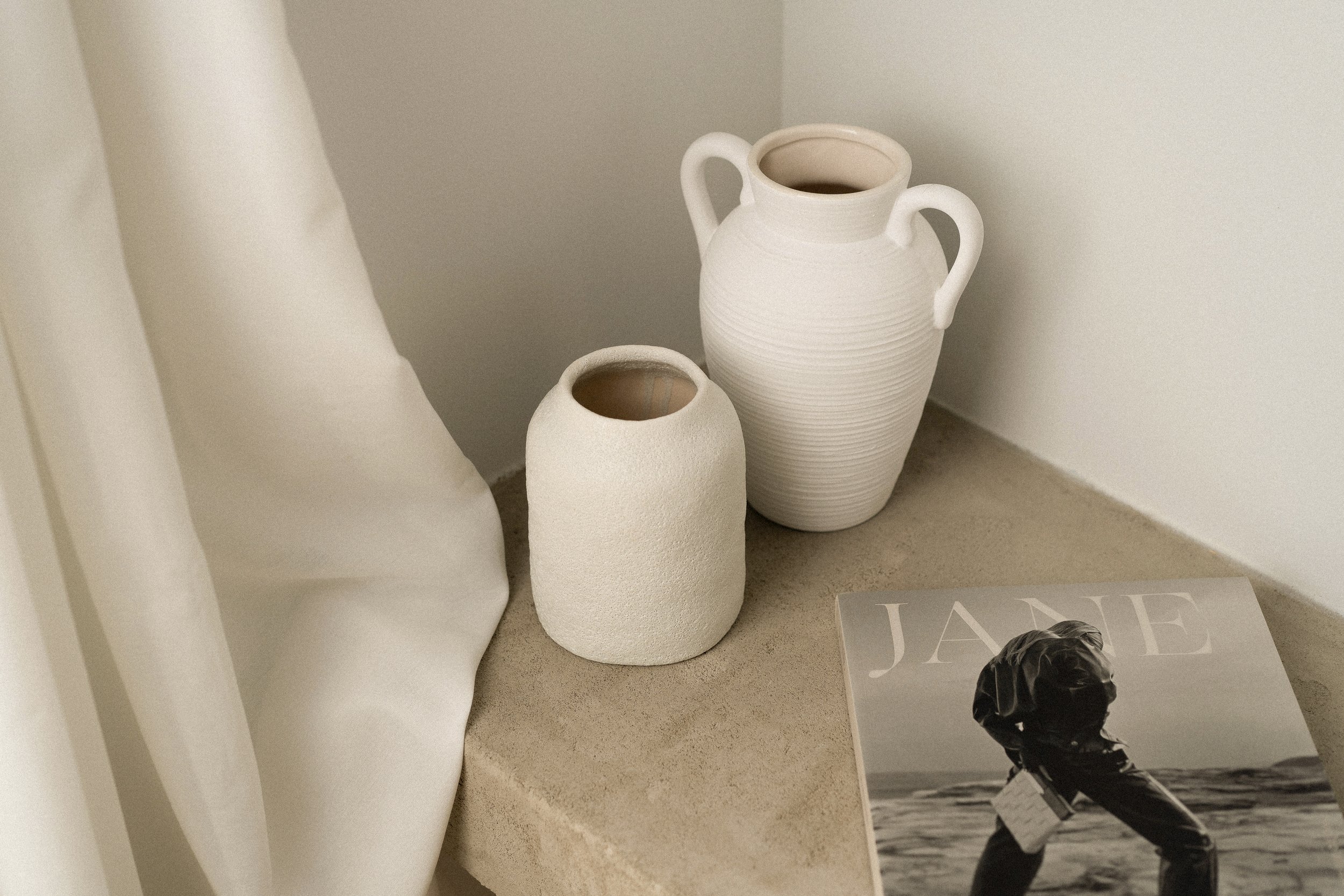 Two white ceramic vases on a stone surface next to a magazine titled "JANE" with a black and white cover image.