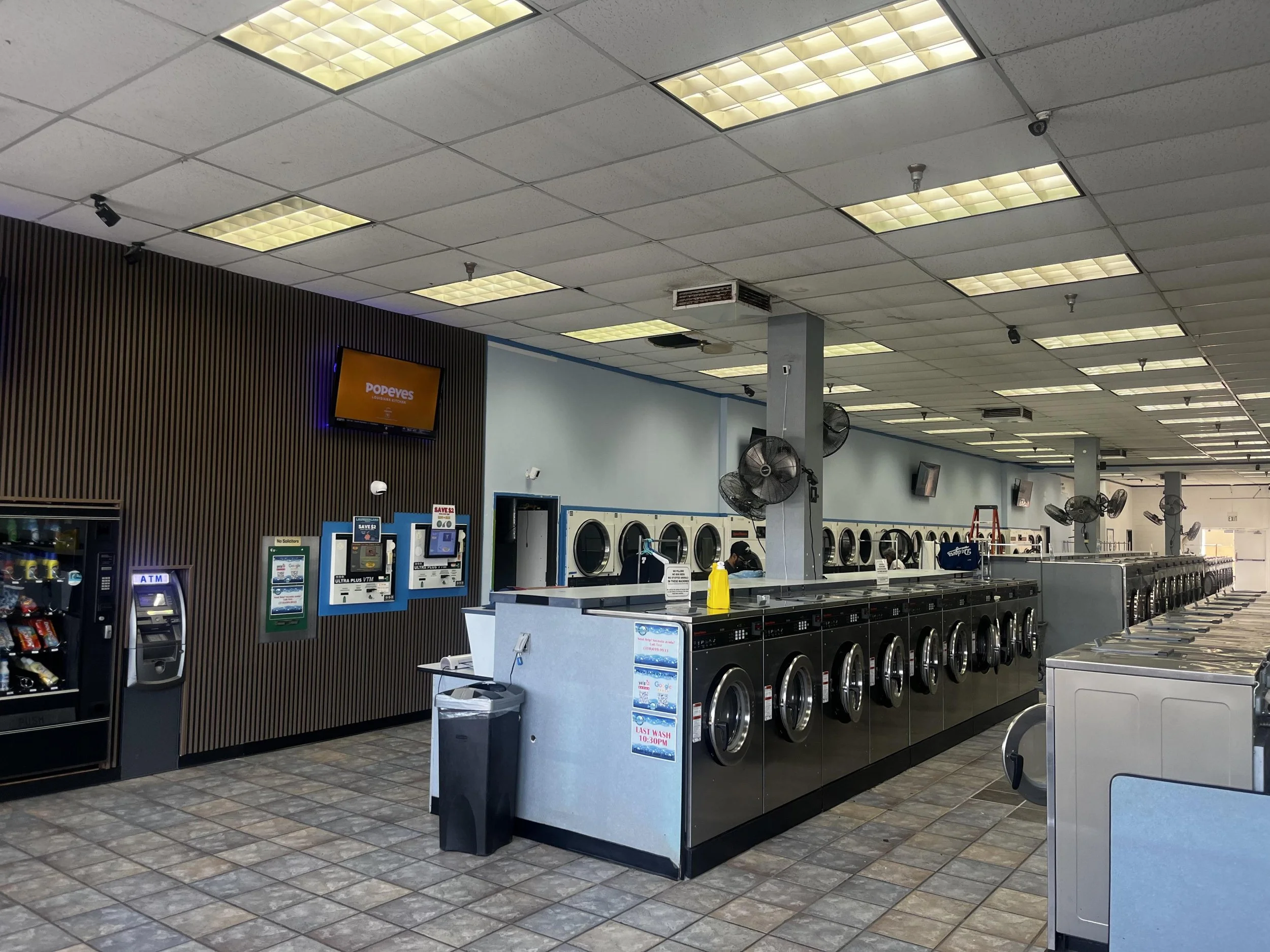 Interior of a laundromat with multiple washing machines and dryers, wall-mounted fans, vending machine, and ATM, with a digital screen on the wall displaying 'Popeyes'
