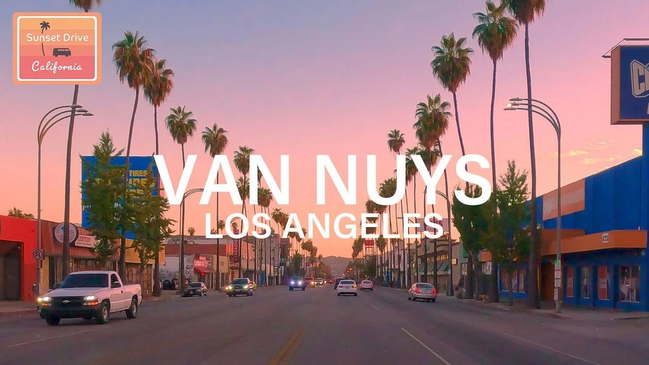 Street view of Van Nuys, Los Angeles during sunset with pink and purple sky, palm trees lining the street, and storefronts on both sides.