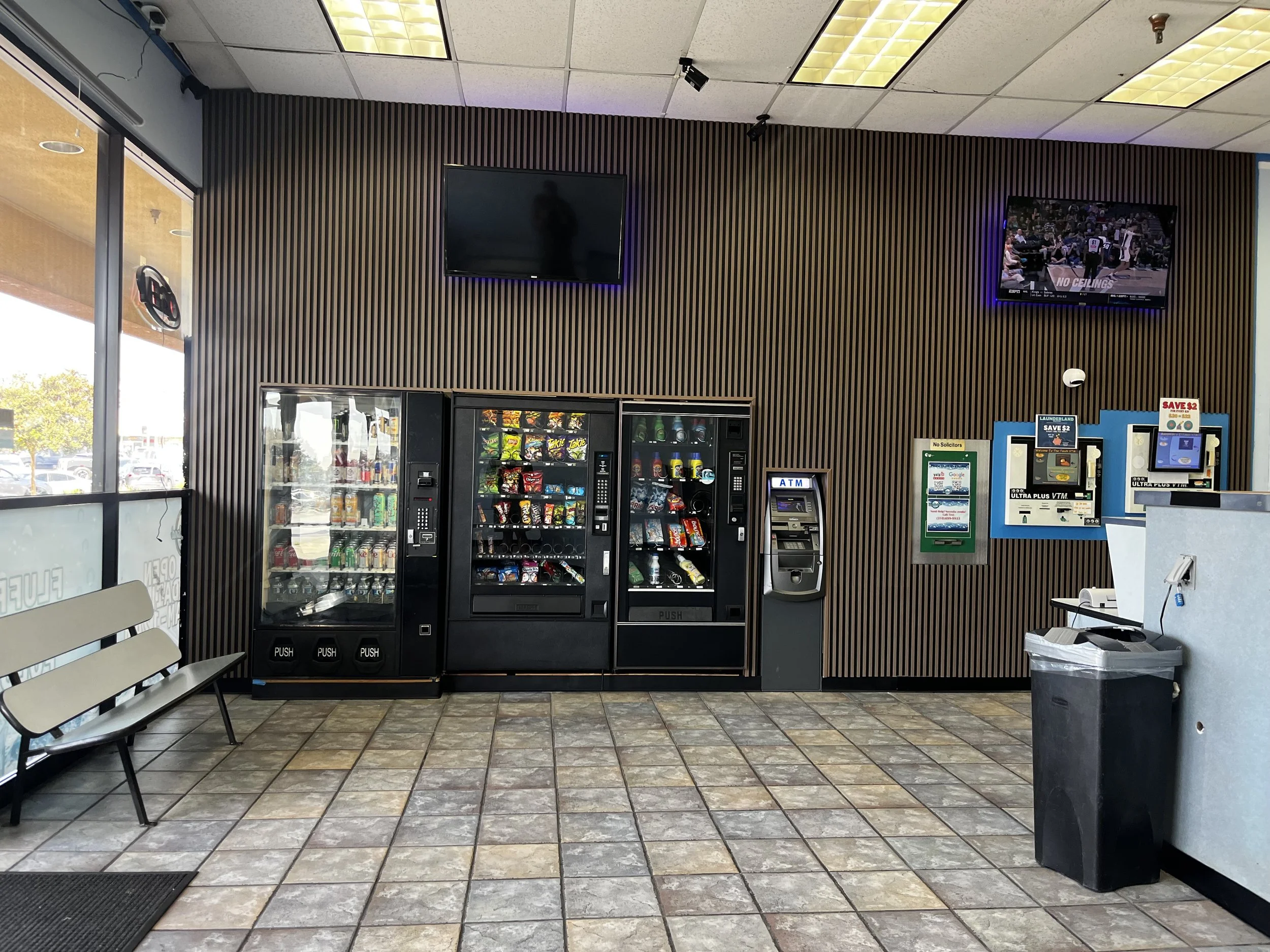 Inside a waiting area with vending machines, a bench on the left side, two wall-mounted televisions, and a wall with a wood slat design. One vending machine is filled with snacks, and a soda machine is beside it. Two ATMs are mounted on the wall, and a trash can is in the foreground on the right side.