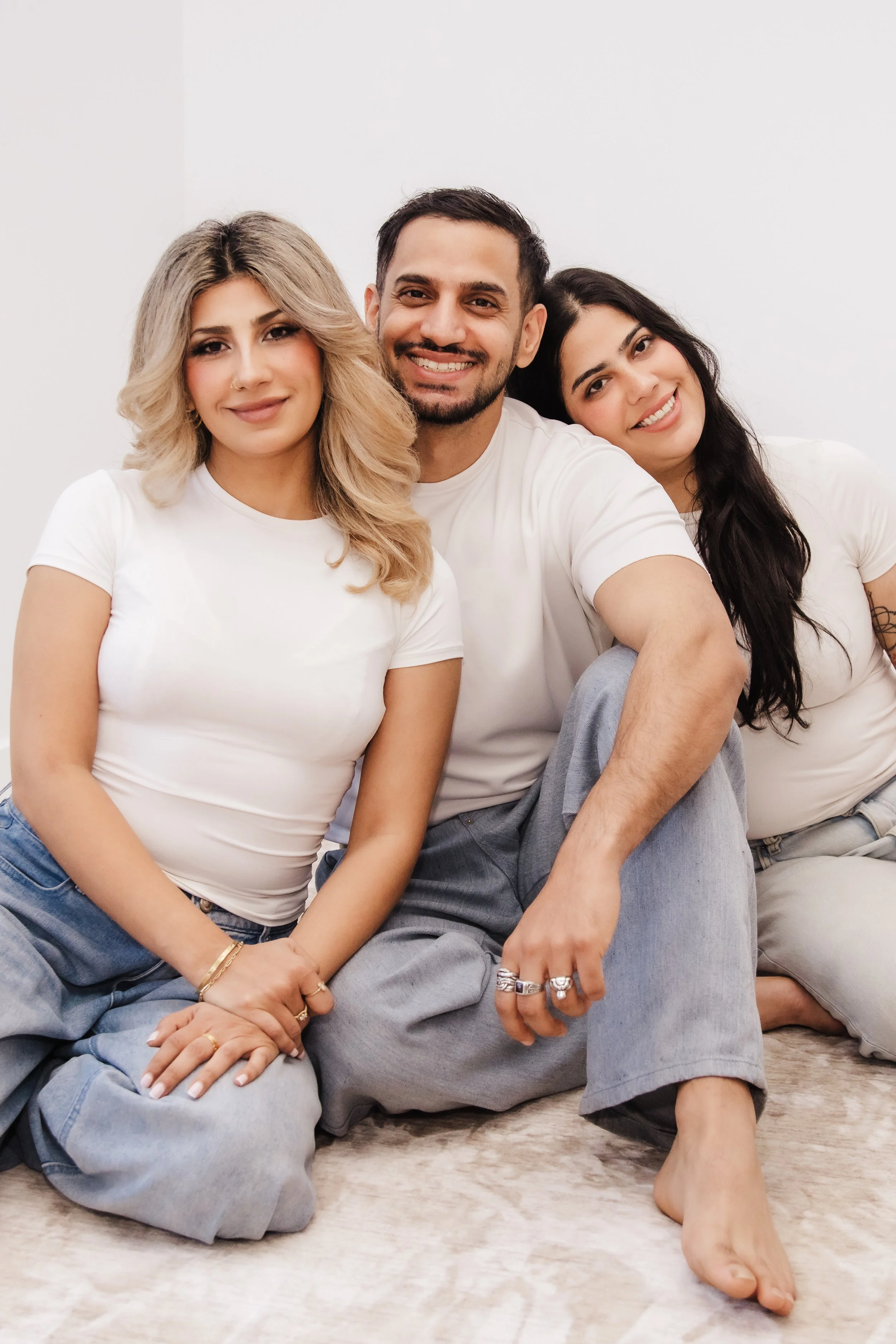 Three people sitting on the floor, smiling, in front of a plain white wall. Two women and one man, casual clothes, rings and jewelry.
