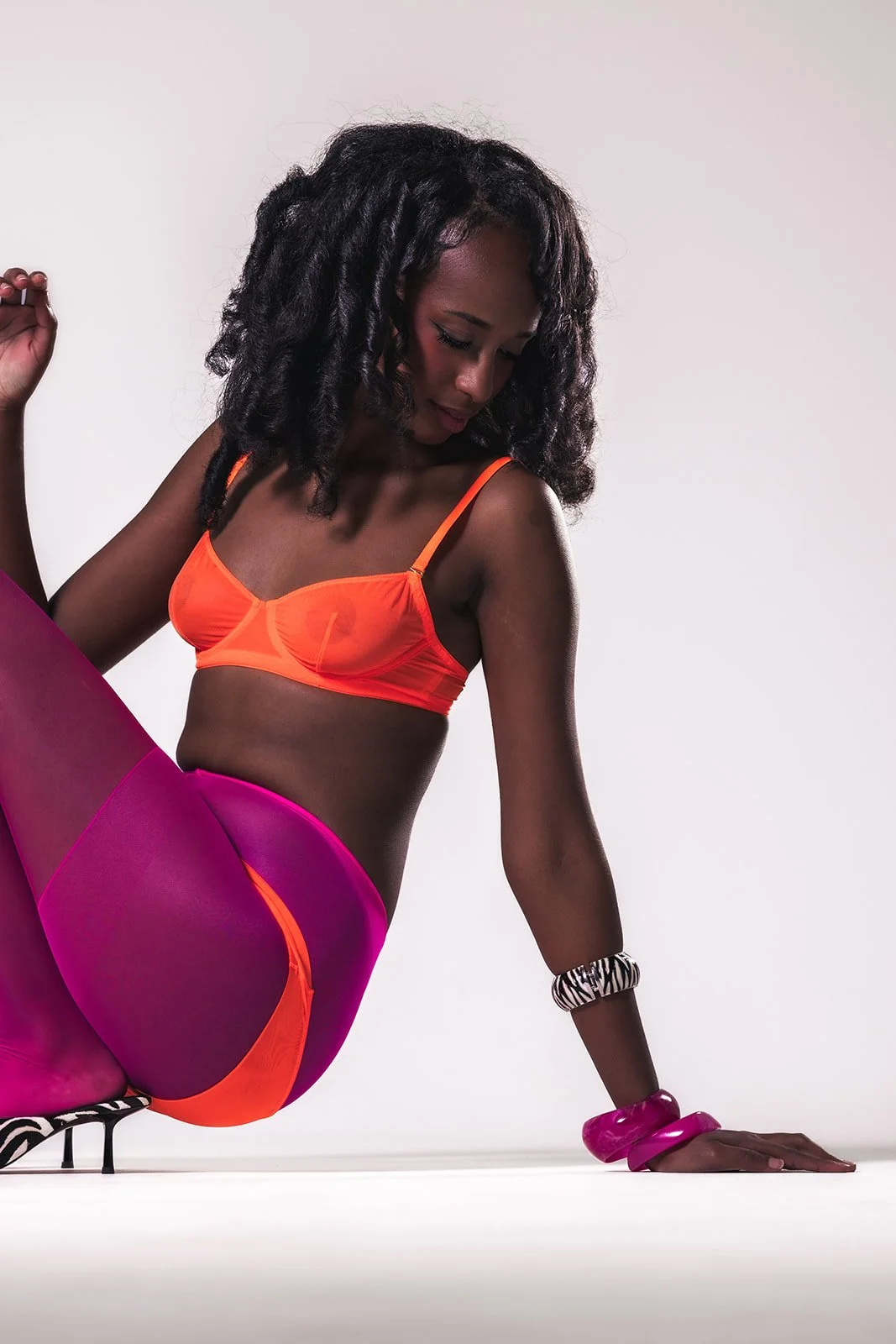 A woman in an orange bra and pink leggings poses on the floor, wearing striped shoes and zebra-print bracelets.