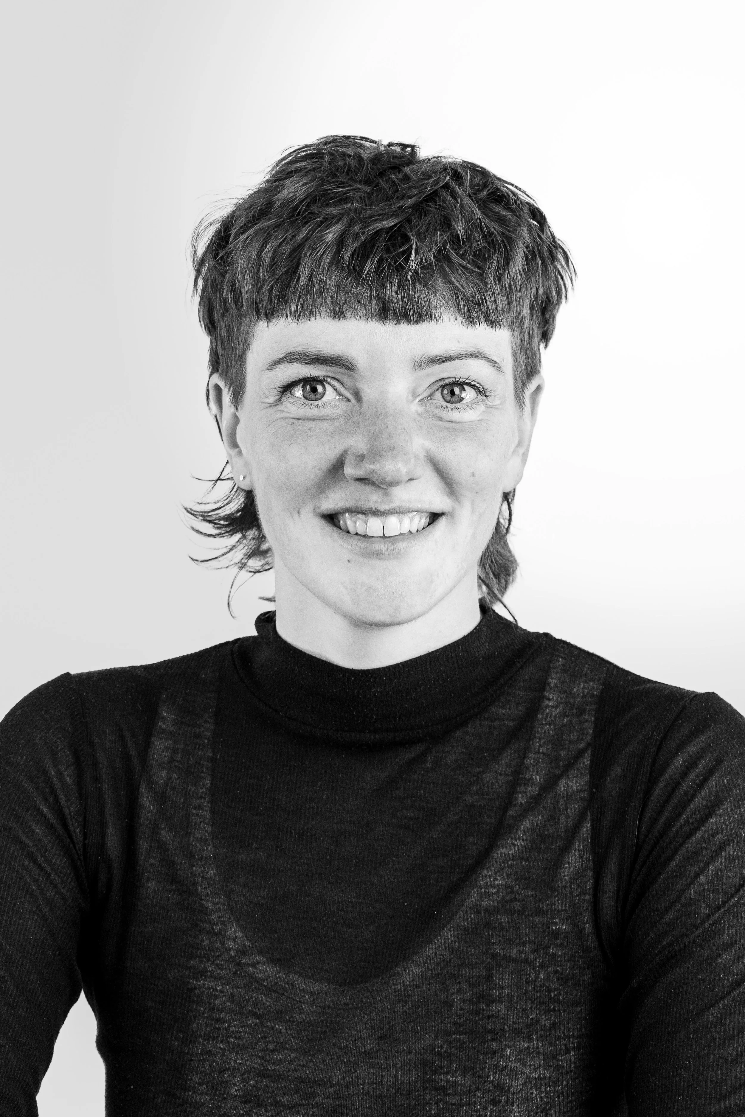 Porträt einer lächelnden Person mit kurzem, lockigem Haar und einem schwarzen Oberteil, vor einem weißen Hintergrund.