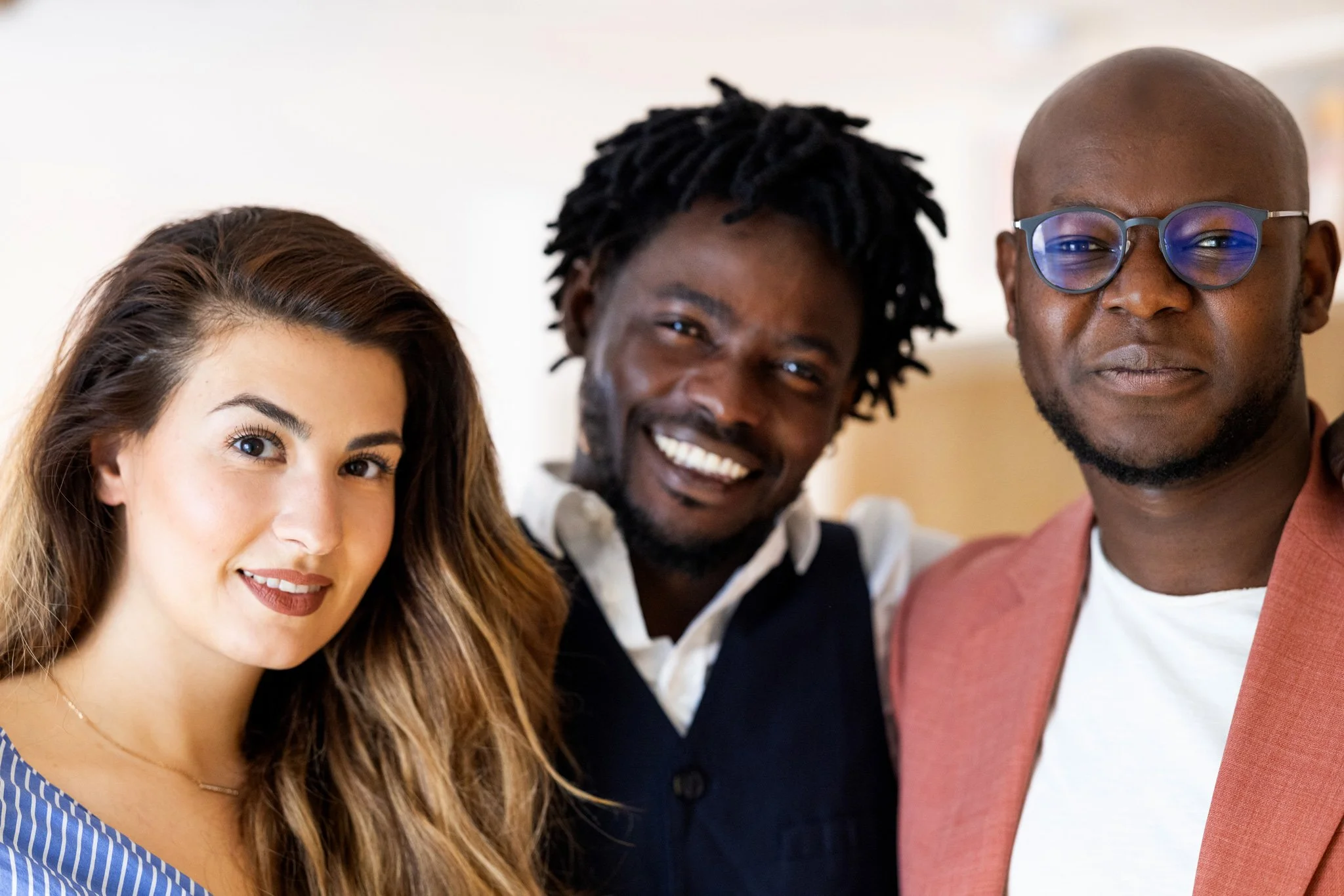 Drei Personen posieren gemeinsam und lächeln in die Kamera. Eine Frau mit langem braunen Haar links, ein Mann mit Dreadlocks in der Mitte, und ein Mann mit Glatze und Brille rechts. Die Stimmung ist freundlich und entspannt.