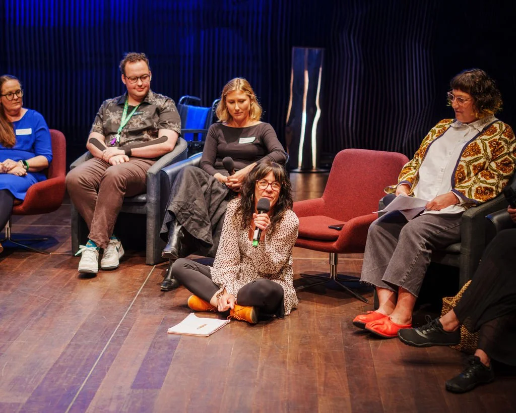 A group of people in discussion. The person with the microphone is sitting ont he floor, while others sit around her on chairs.