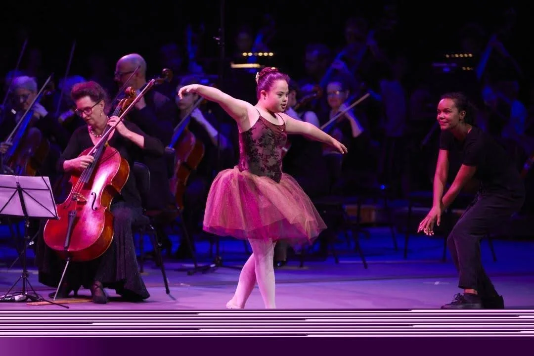 A ballet dancer on stage in a tutu with an orchestra in the background under stage lighting.