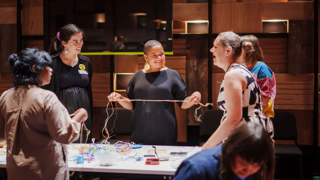 A group of people smiling and chatting with each other. The woman in the middle is holding out a long piece of raffia, as if to begin weaving. There is coloured raffia on the table in front of them.