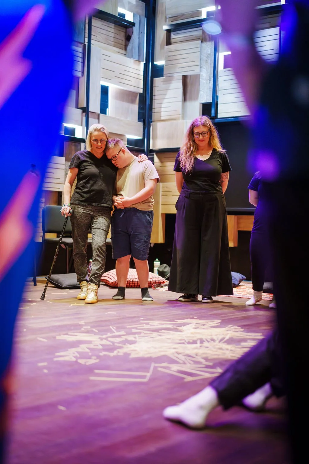 Three people look toward the ground where there are hundreds of strewn wooden sticks. One woman using a cane puts her arm around a young man, while he rests his head on her shoulder.
