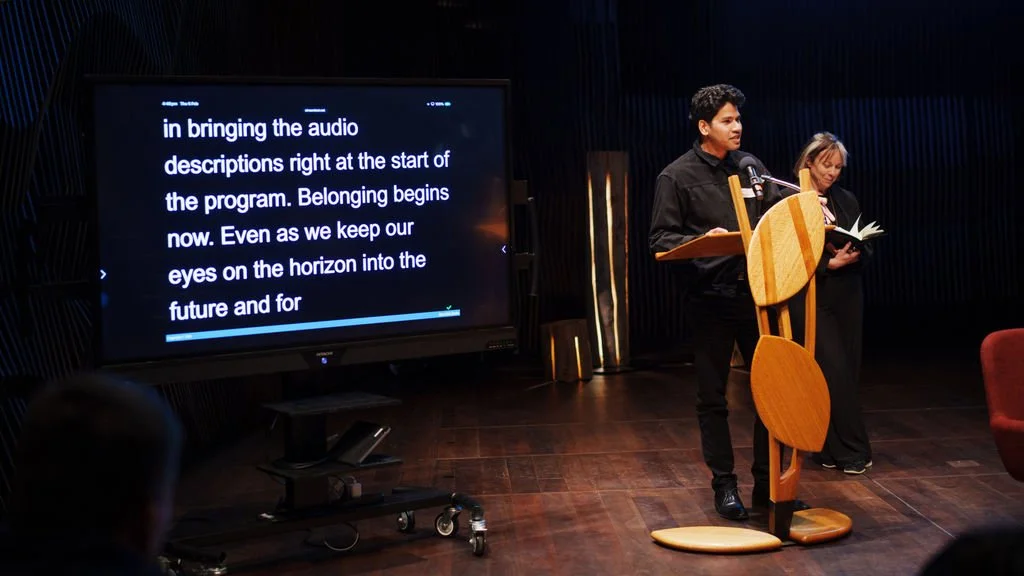 A man speaking into a microphone at a decorative wooden lecturn. There is a tv monitor with live captioning on screen in the foreground.