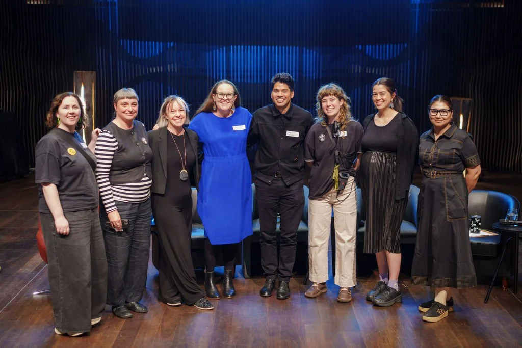 A group of 8 people stand in a row, smiling at the camera. Morwenna is in the middle wearing a bright blue dress.
