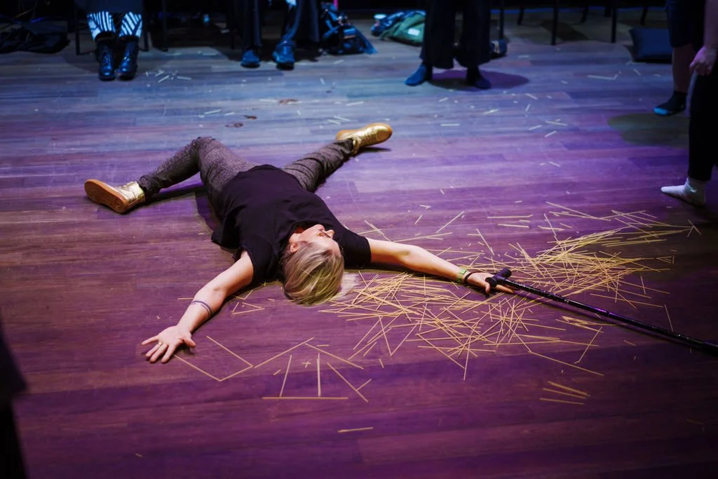 A person lays spread out on the floor - all limbs outstretched. In her right hand is her cane, and underneath here are hundreds of tiny sticks that looks like dried spaghetti