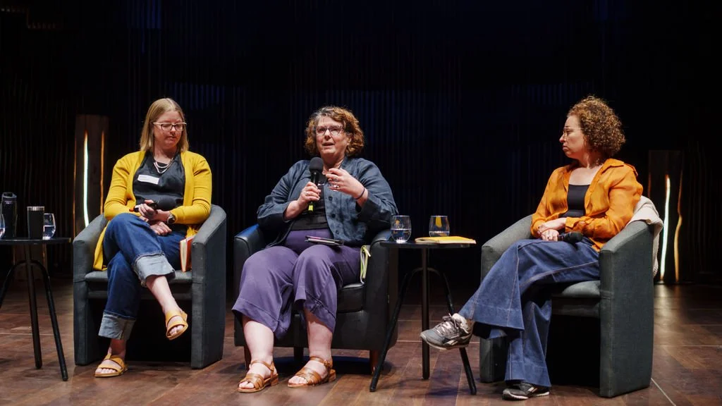 Three women sitting on stage for a panel discussion. The woman in the middle is holding up the microphone and the other two women are looking toward her.