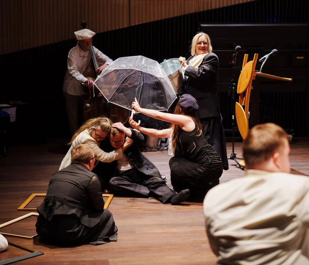 On a stage, performers enact a dramatic scene: several people crouch together under a clear umbrella while another stands holding a metal bucket above them.