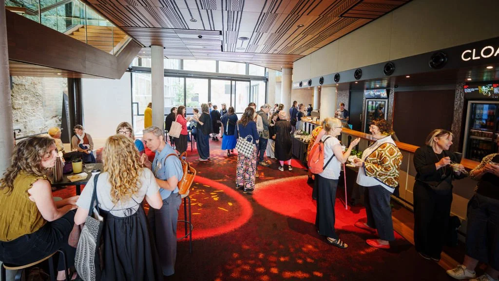 A bright foyer space shows people mingling and chatting, with small groups gathered around high tables and a bar counter. Natural light streams through large windows, creating a warm and lively atmosphere.