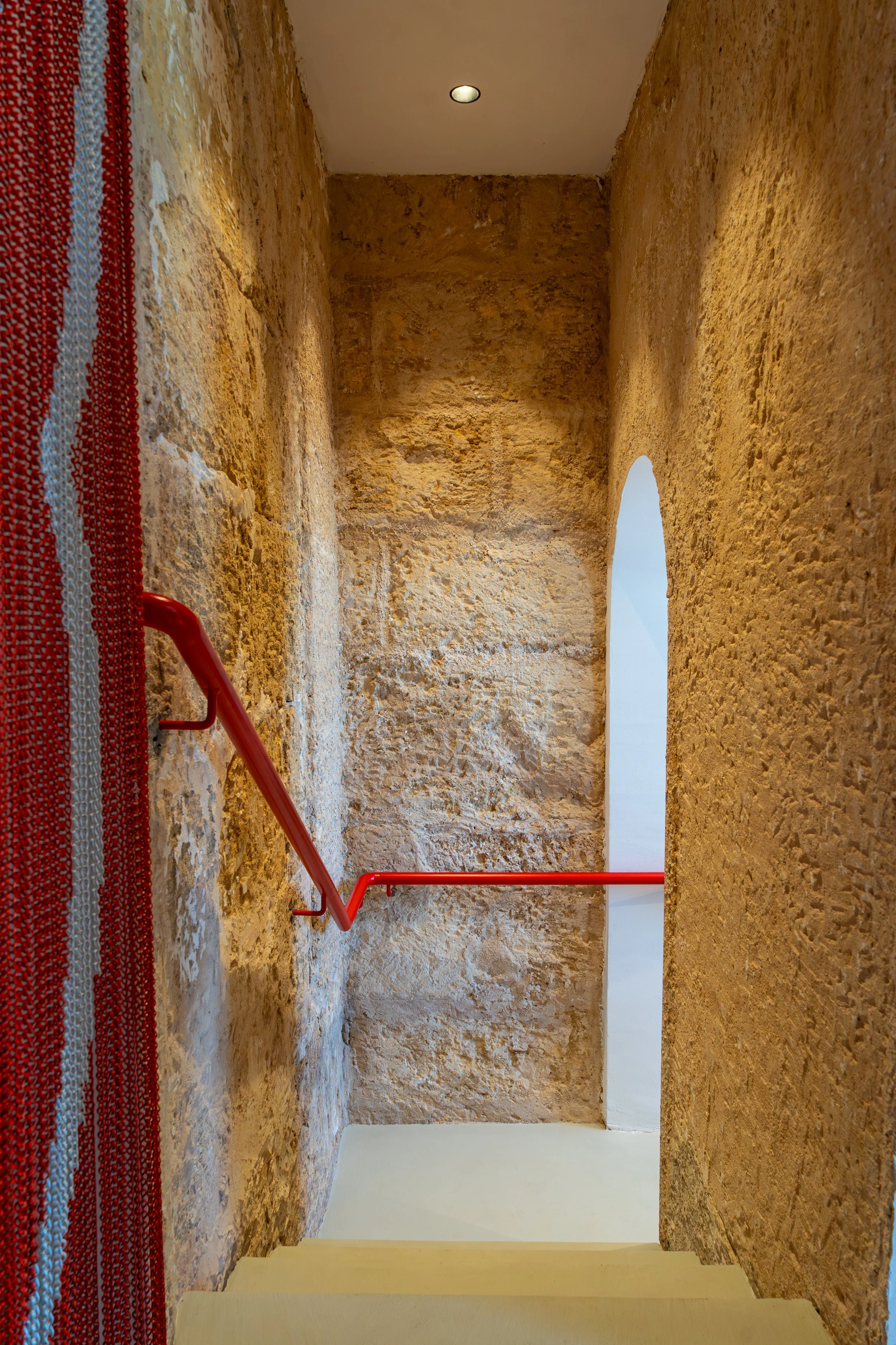 Escalera interior con paredes de piedra, barandilla roja, alfombra de colores en un lado y arco en la pared al fondo.