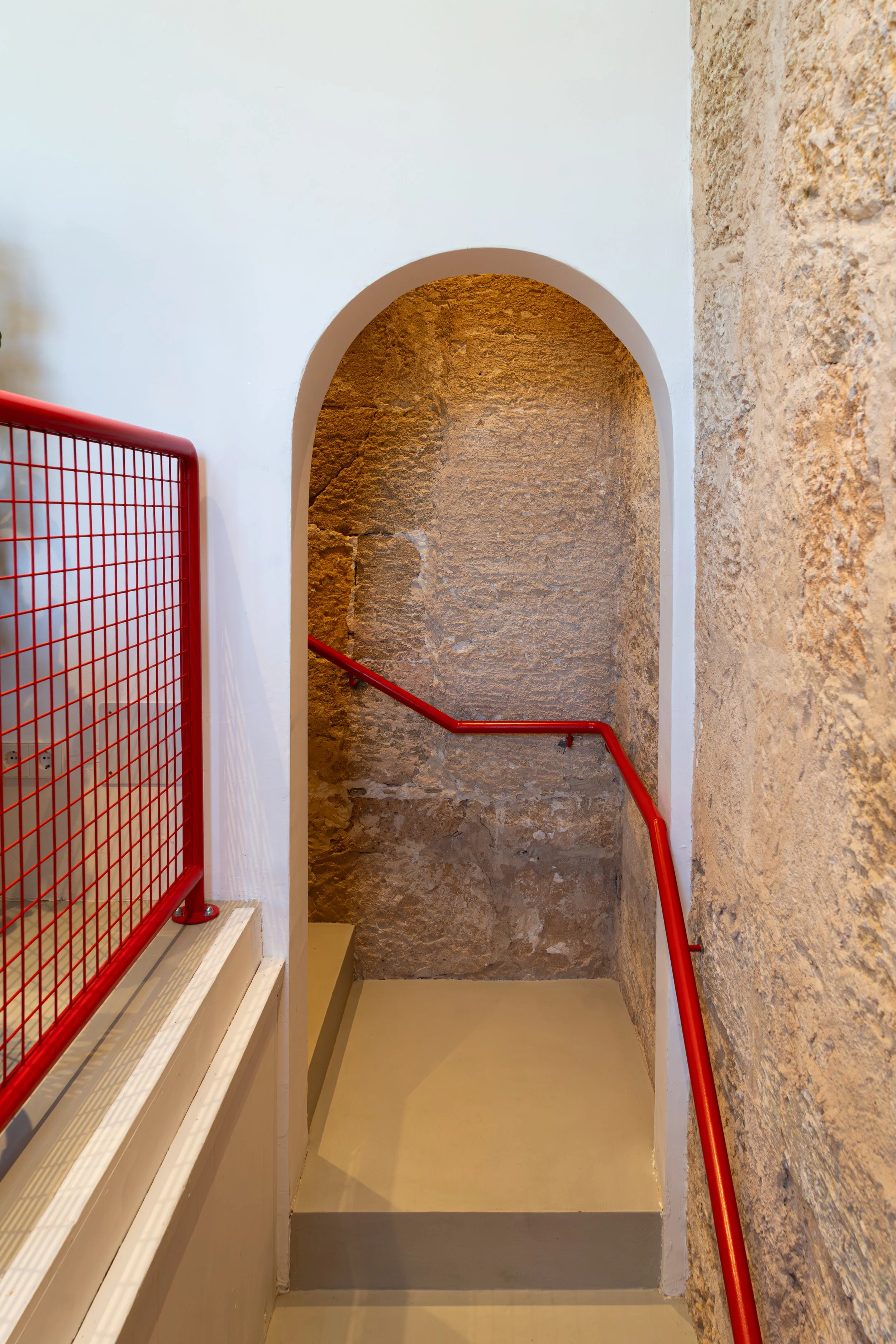 Escalera con pasamanos rojo en un interior con paredes de piedra y arco.