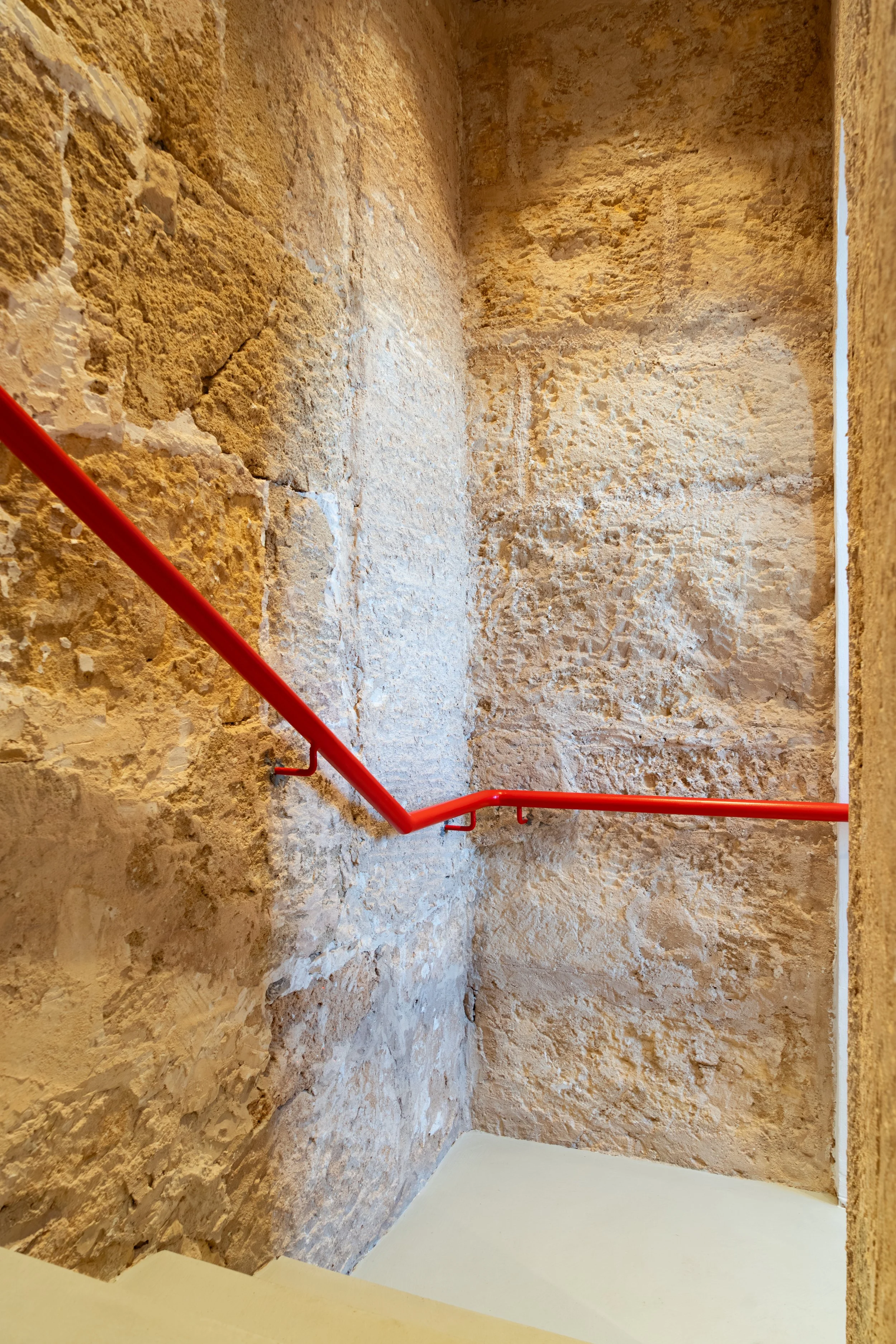Escalera con barandilla roja en un espacio con paredes de piedra y un suelo beige.