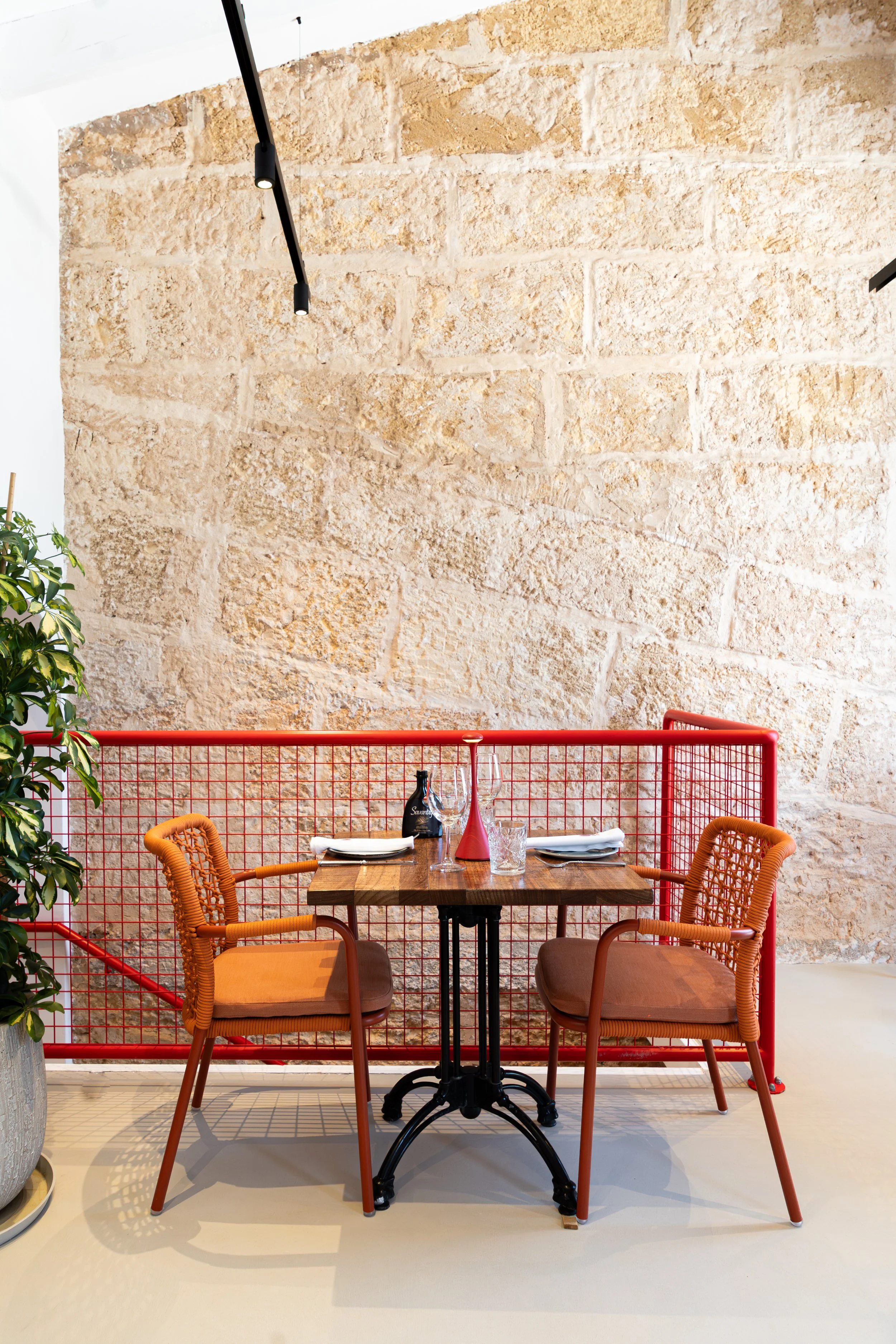 Mesa de comedor rústica con dos sillas de madera y cojines naranjas, decorada con una botella de vino, copas y un florero rosado, en un espacio cerrado con paredes de piedra y una división metálica roja.