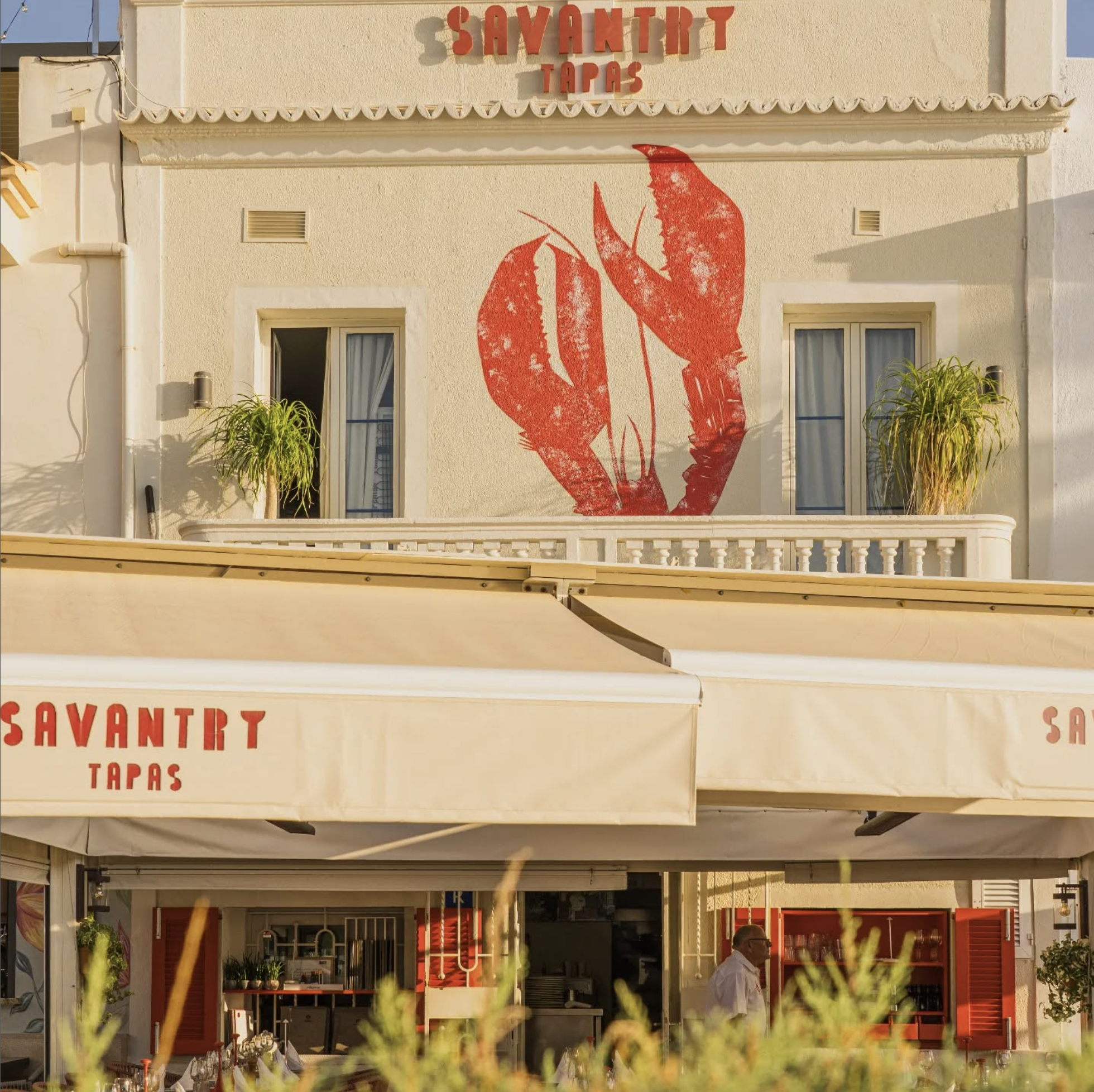 Fachada de un restaurante llamado "Savantry Tapas" con un mural de una langosta en la pared y dos ventanales con cortinas blancas, así como plantas en la terraza y un toldo en la parte inferior
