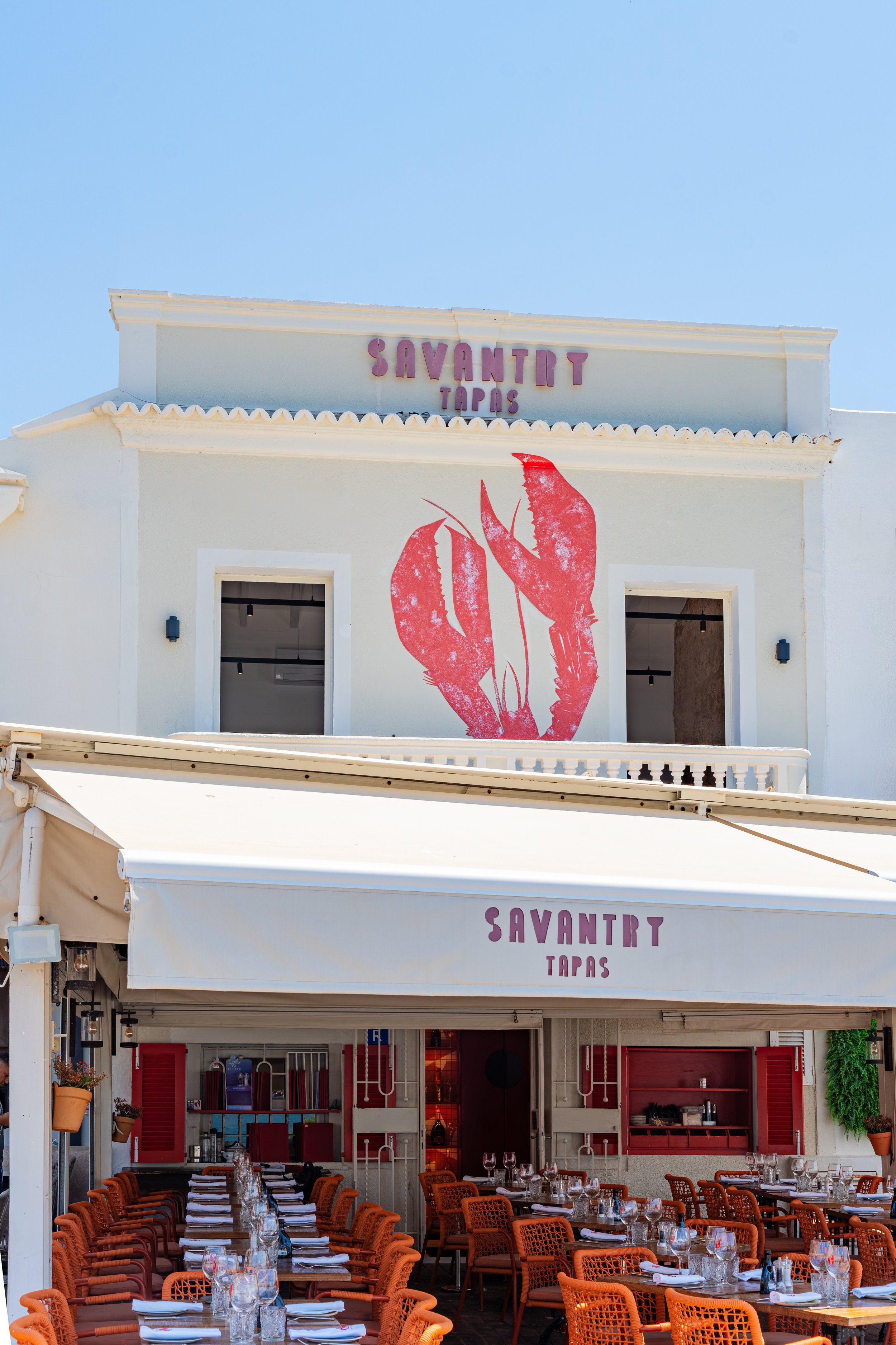 Fachada de un restaurante llamado Savanty Tapas con un gran logotipo de una langosta en la pared. En la parte inferior, una terraza al aire libre con mesas y sillas listas para clientes.