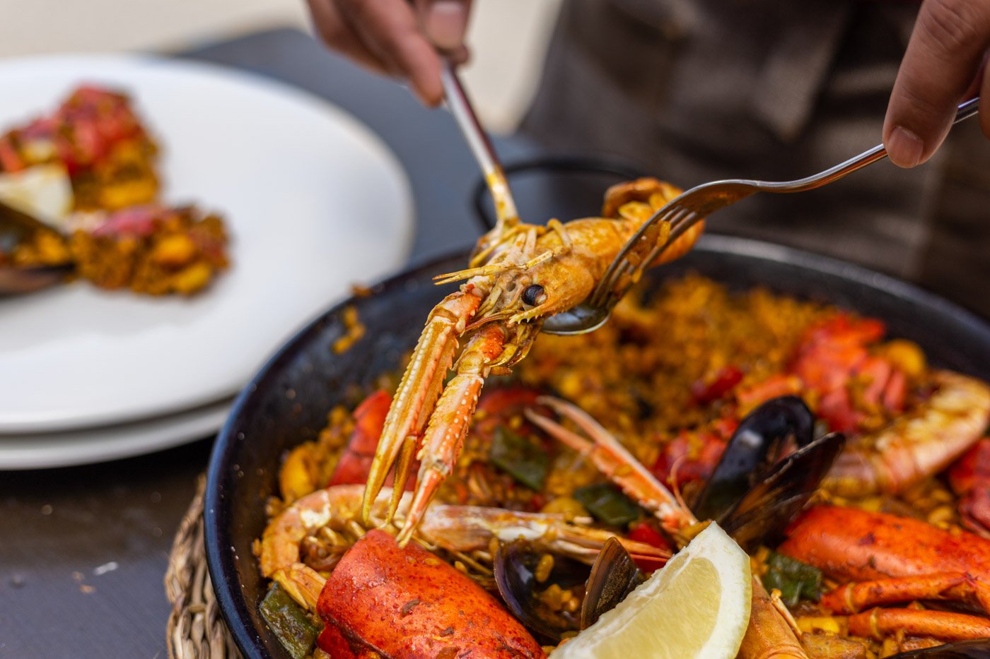 Persona sirviendo mariscos, incluyendo langosta y mejillones, de un plato grande con limón en una mesa.