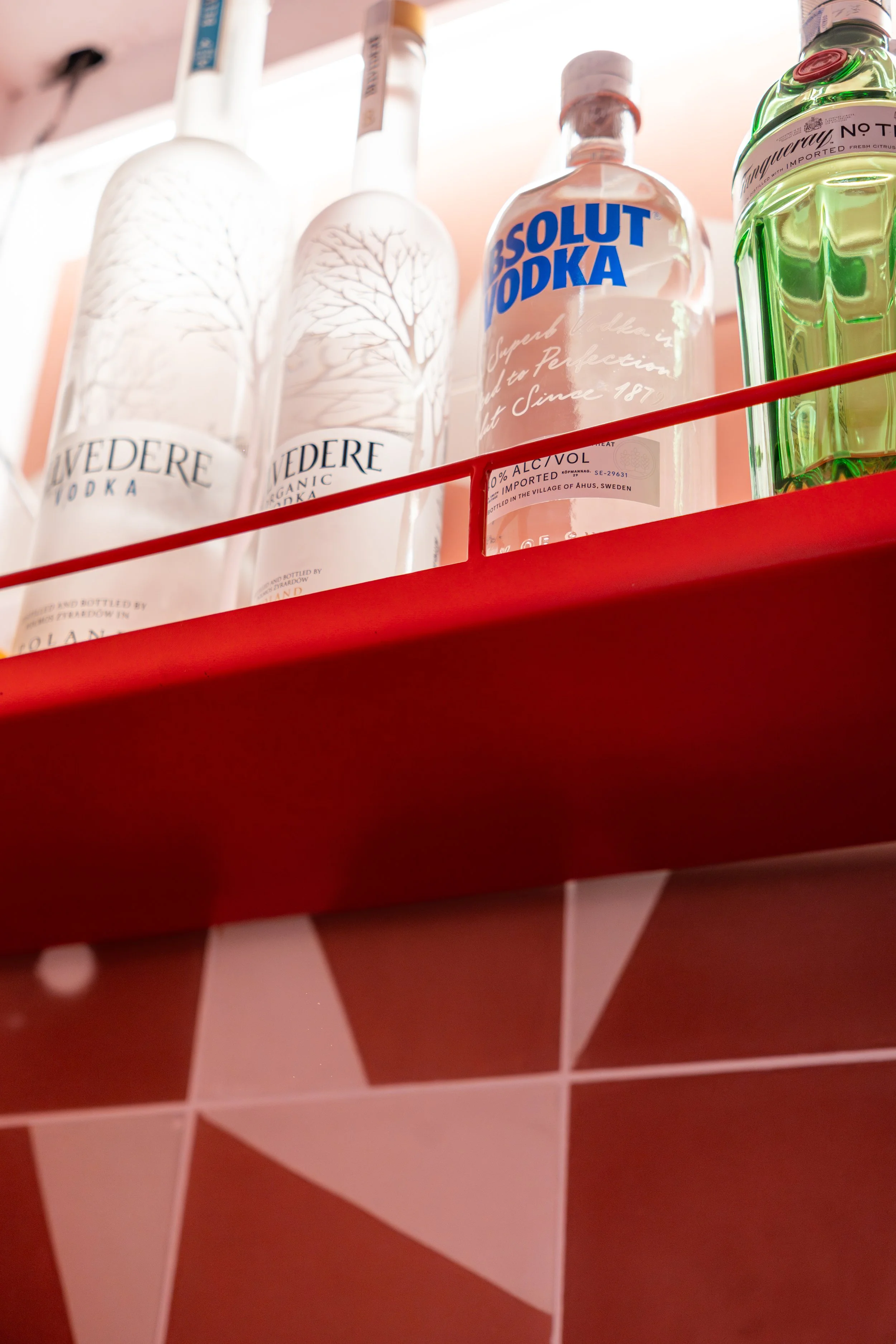 Botellas de vodka en una estantería, incluyendo marcas como Vedre, Absolut y Tanqueray, en un fondo con azulejos rosados y una borde de estante rojo.
