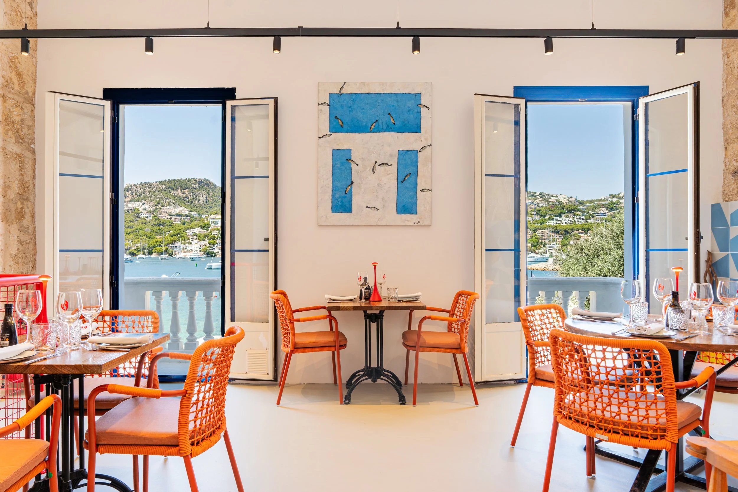 Restaurante con vista al mar, mesas con cubiertos y copas, sillones naranjas, puertas abiertas hacia un paisaje oceánico y colinas