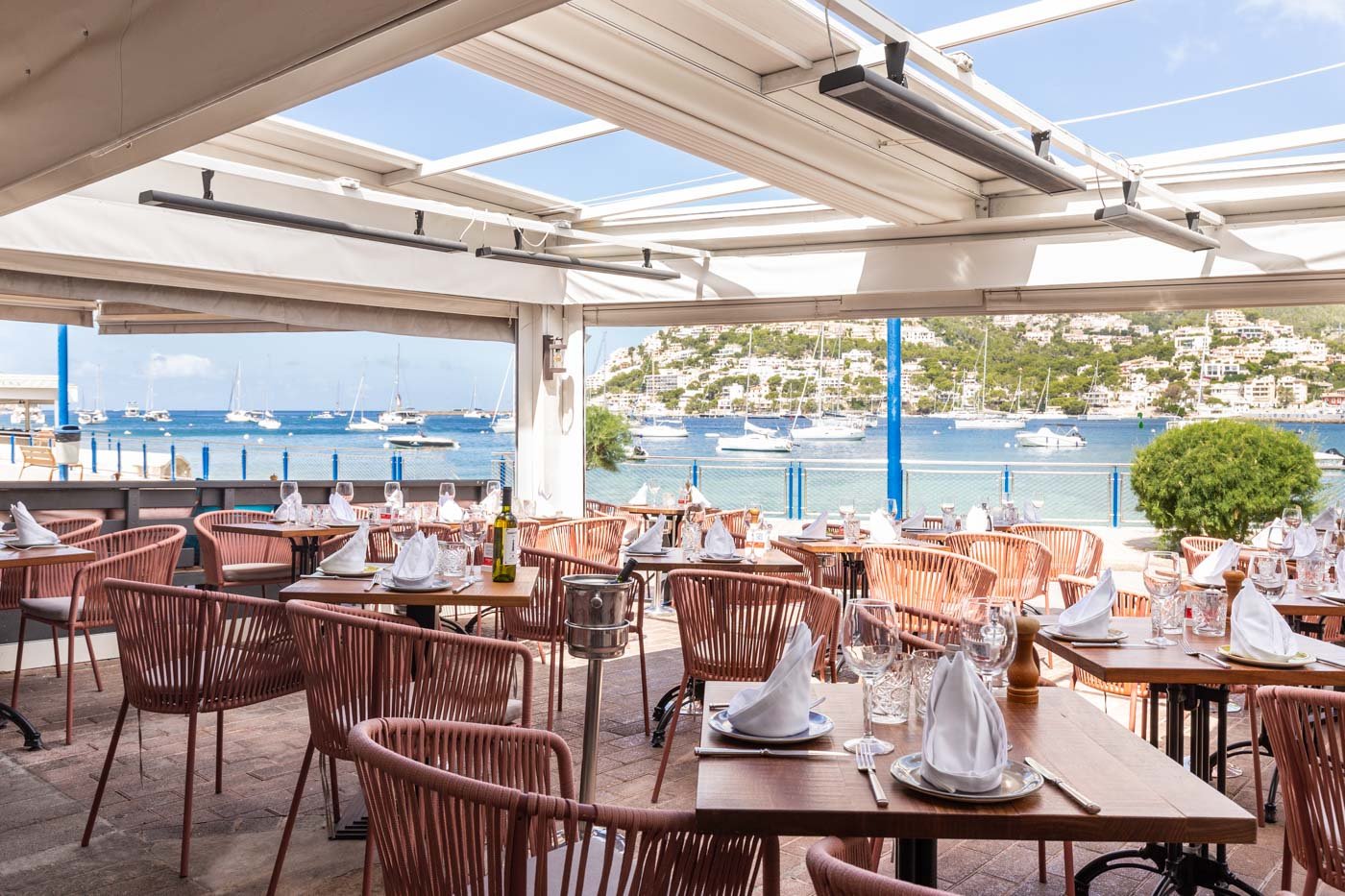 Restaurante con vista a un puerto con barcos y casas en la colina, mesas con manteles blancos y utensilios preparados para comer.