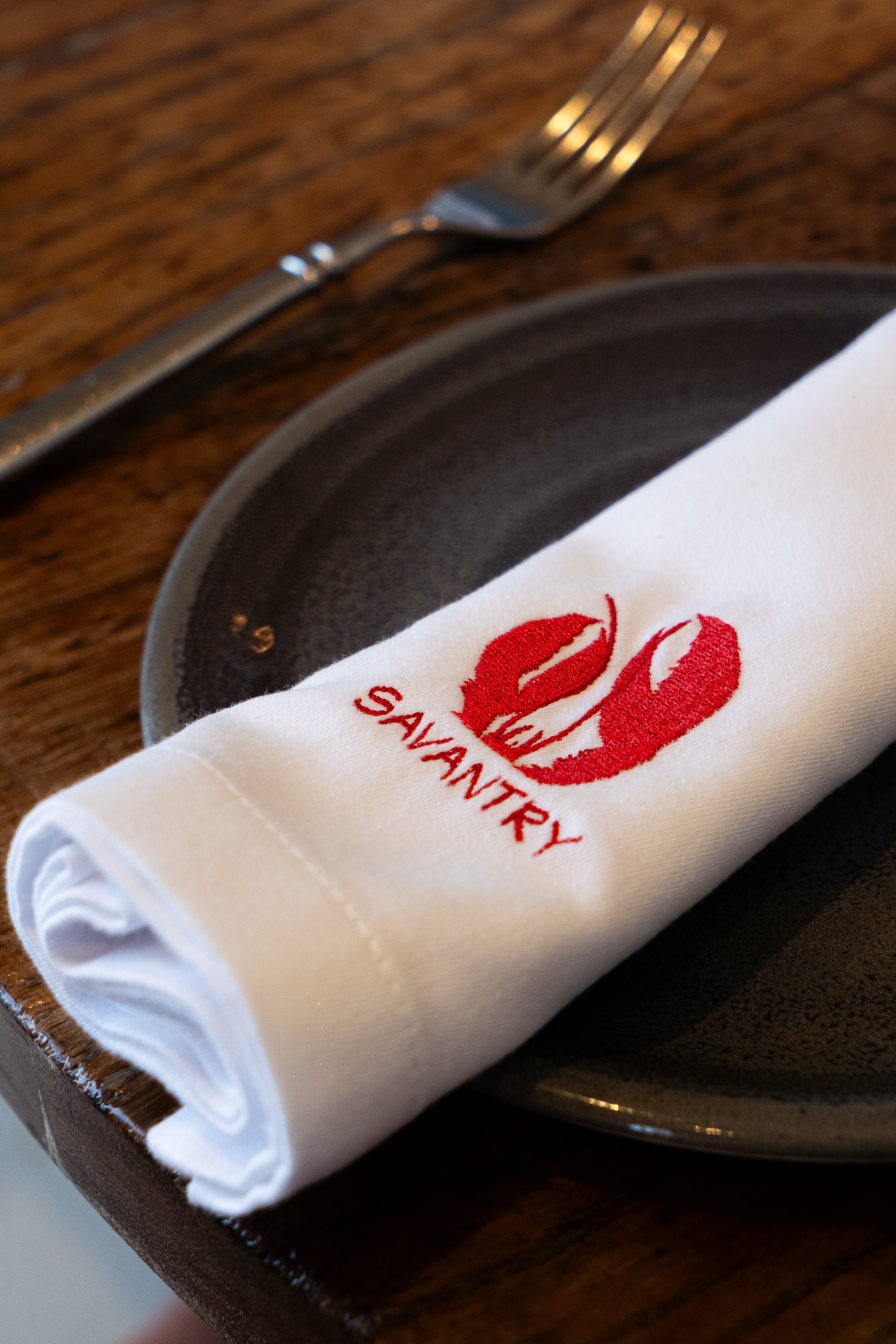 Servilleta blanca con bordado rojo de labios y la palabra 'Salvatory', colocada sobre un plato negro en una mesa de madera junto a un tenedor.