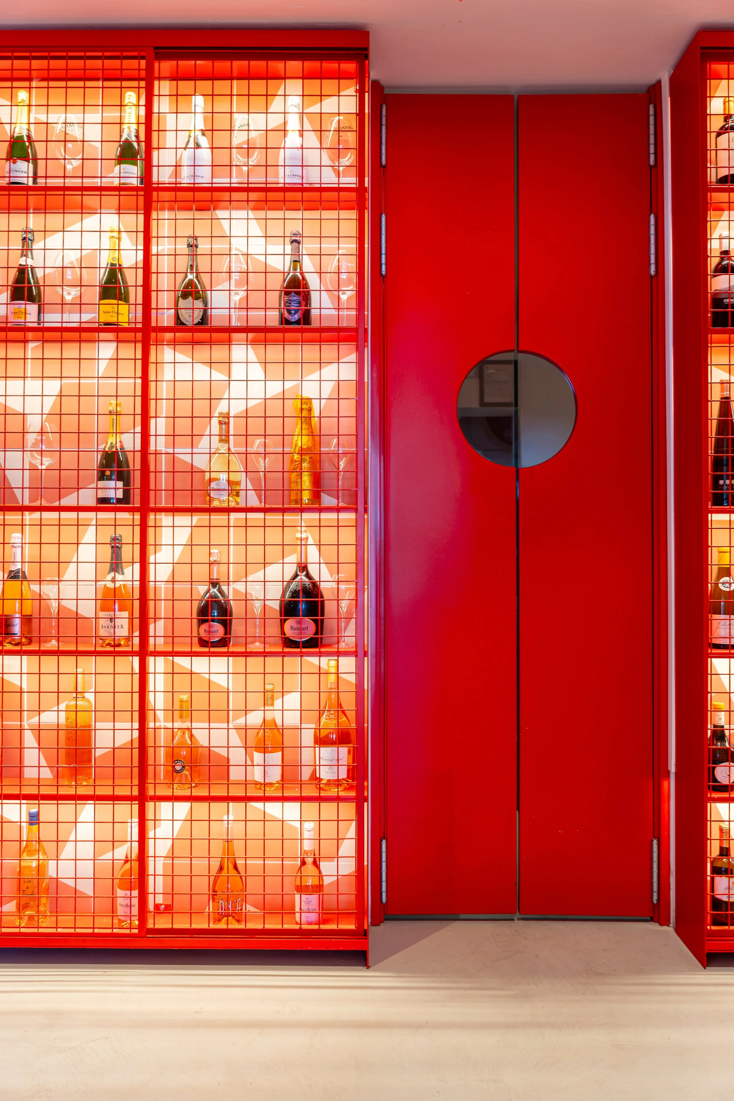 Estantería de botellas de champagne con una pared de fondo de color naranja y una puerta roja con una ventana circular.
