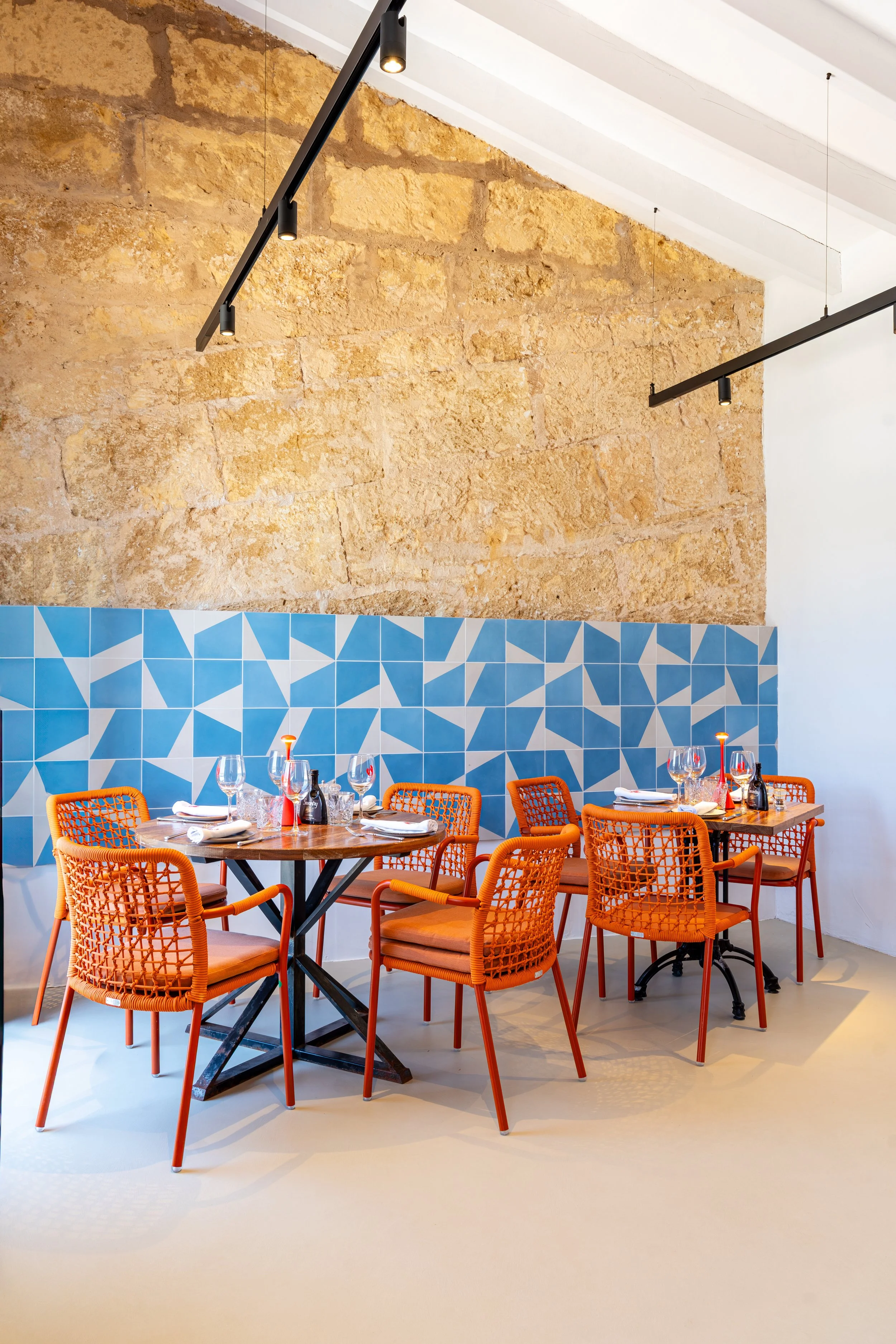 Mesa de comedor en un restaurante con cuatro sillas naranjas de mimbre y piso beige, con paredes de piedra y azulejos azules con patrón geométrico, y dos lámparas negras suspendidas en el techo.
