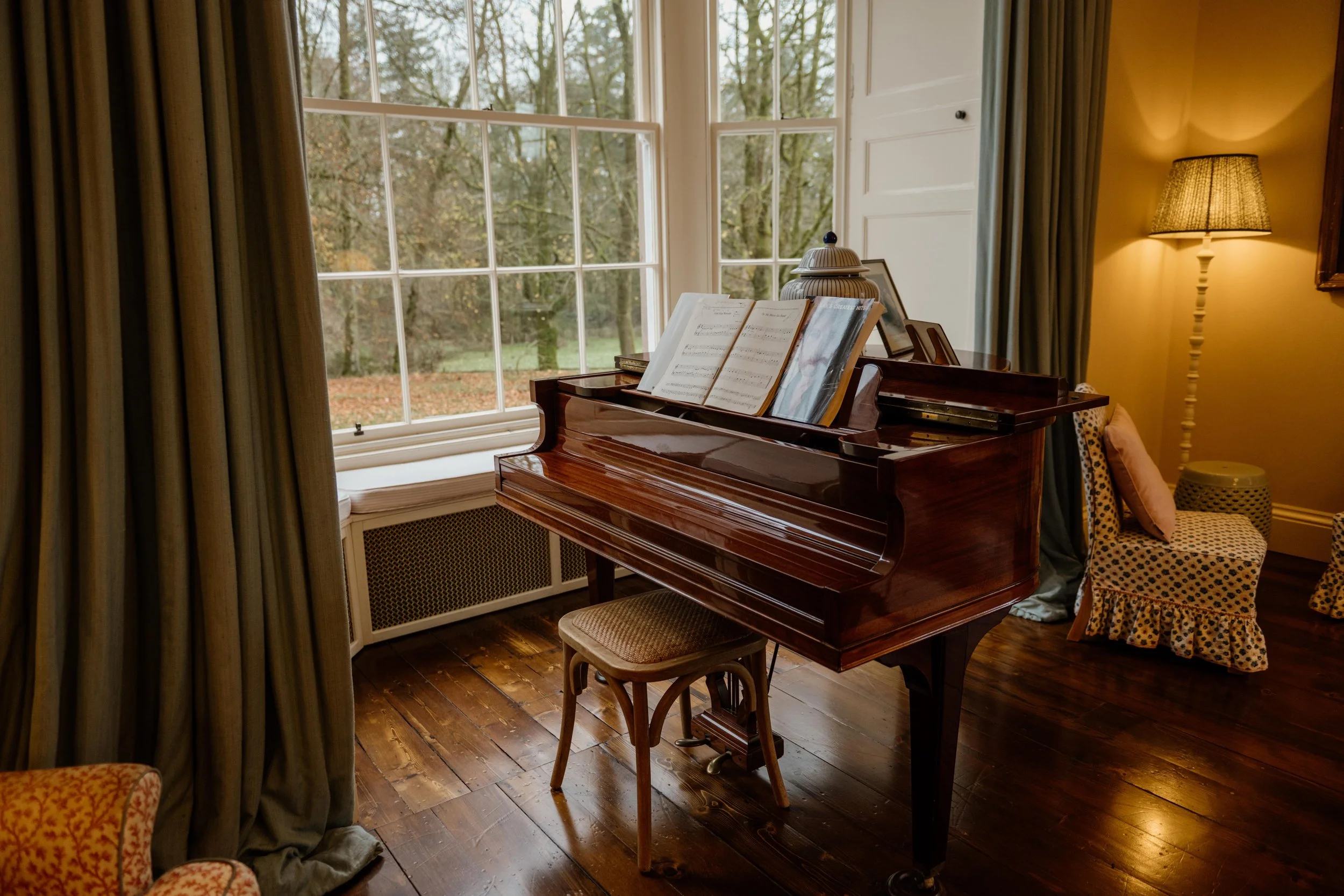 A cozy room with a grand piano next to large windows overlooking a wooded landscape, with a chair and a stool nearby, warm lighting from a lamp, and curtains framing the scene.
