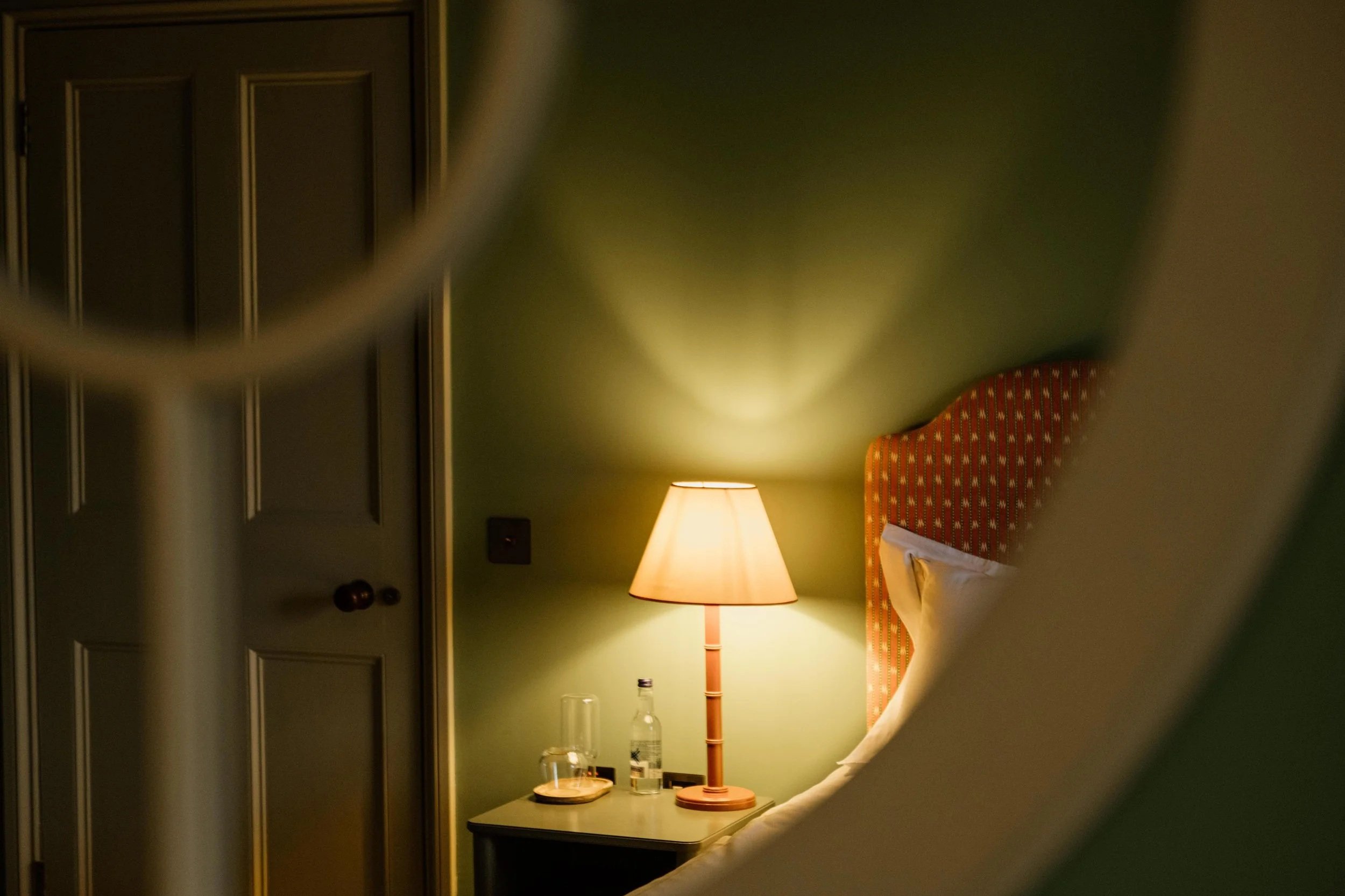 A bedside scene viewed through a partially obscuring object, showing a lamp on a nightstand, a bottle, a glass, and part of a bed with a headboard.