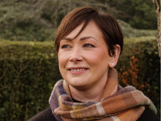 A woman with short brown hair wearing a colorful plaid scarf and a black sweater, standing outdoors near a tree with a background of greenery and a cloudy sky.