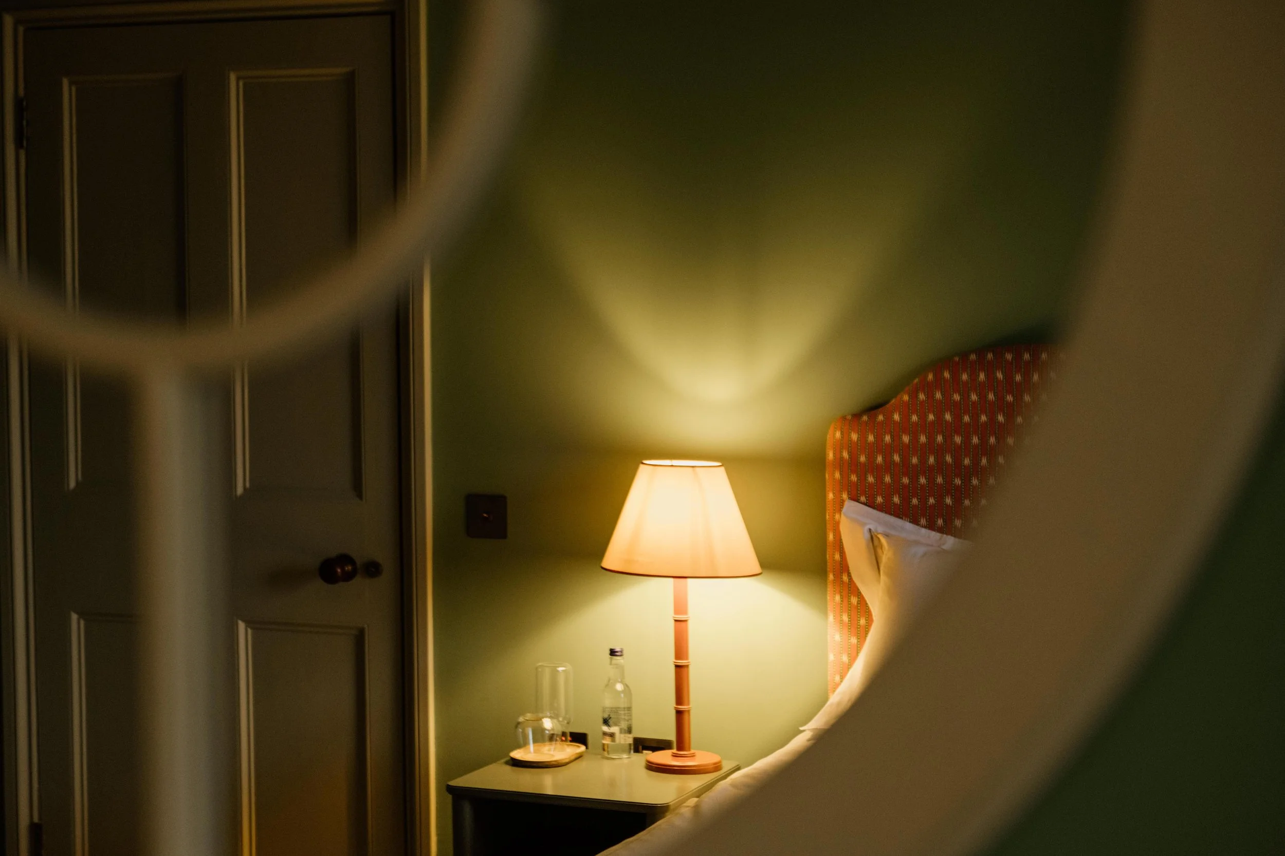 Nightstand with a table lamp, water bottles, and glass, a bed with a red patterned headboard, and closed closet door in a dimly lit hotel room.