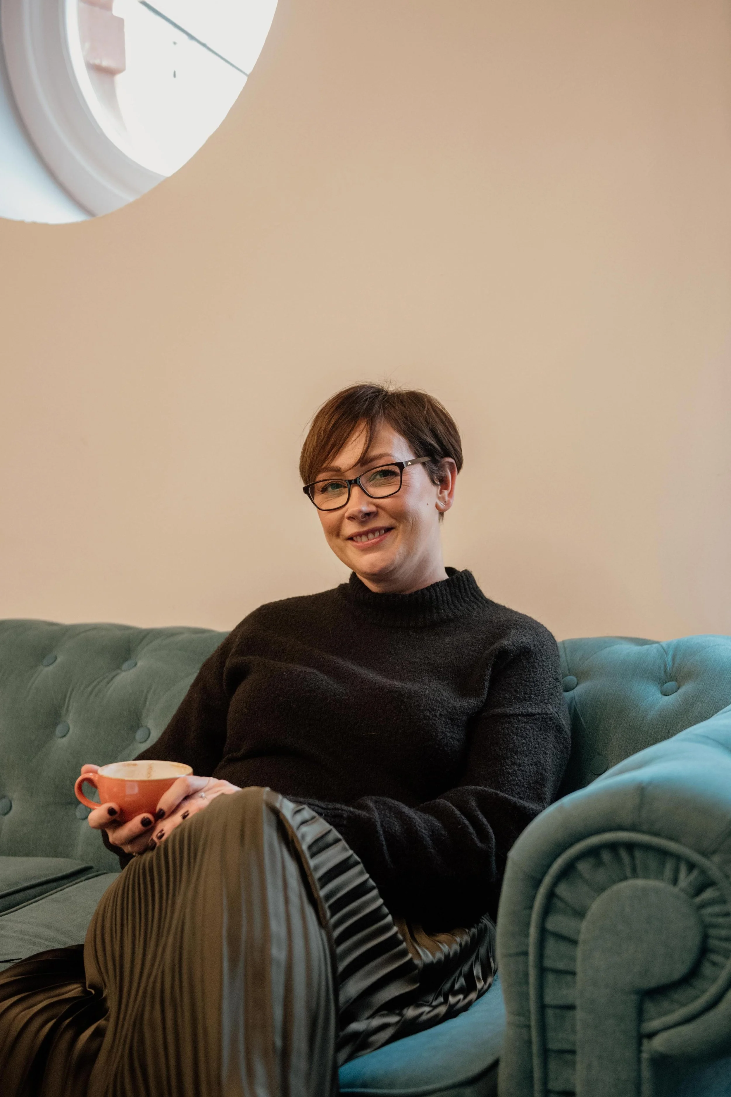 A woman with short brown hair, glasses, and a black sweater sitting on a teal tufted couch, smiling, holding a pink mug, in a room with a beige wall, and a window in the background.