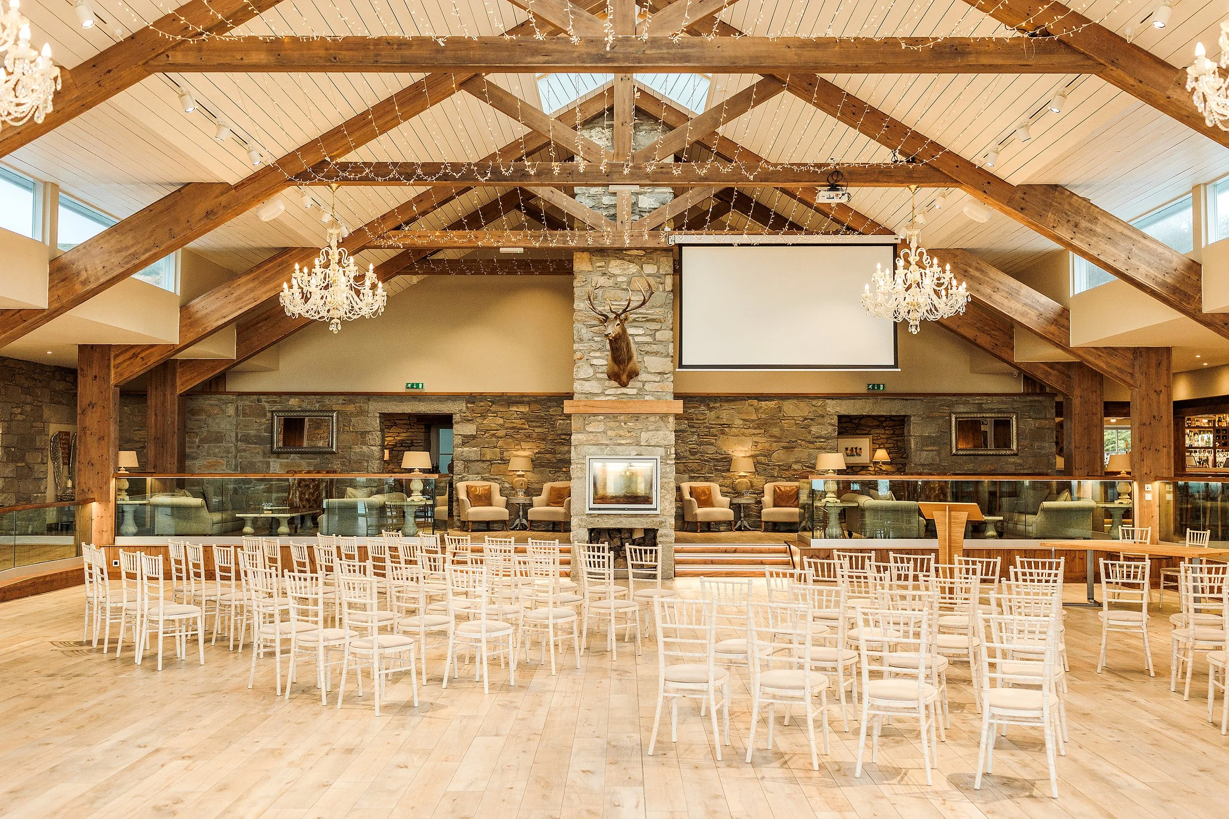 An indoor event space with wooden beams, chandeliers, and a ceiling decorated with string lights. There is a stone fireplace with a mounted deer head and a projection screen on the wall. Rows of white chairs are arranged facing the front, with additional seating areas visible.