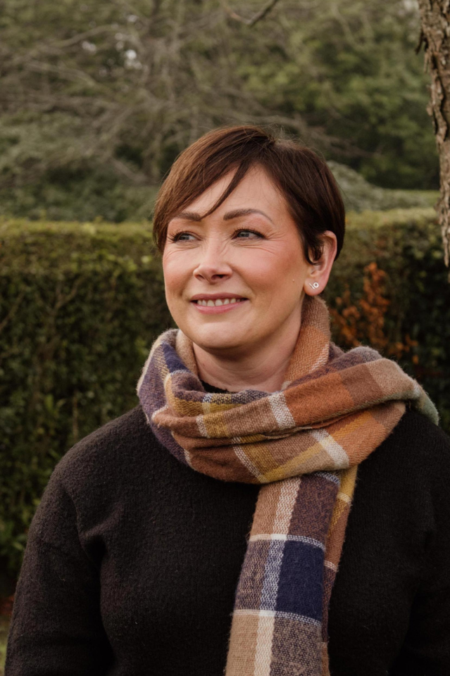 A woman with short brown hair wearing a colorful plaid scarf and a black sweater, standing outdoors near a tree with a background of greenery and a cloudy sky.