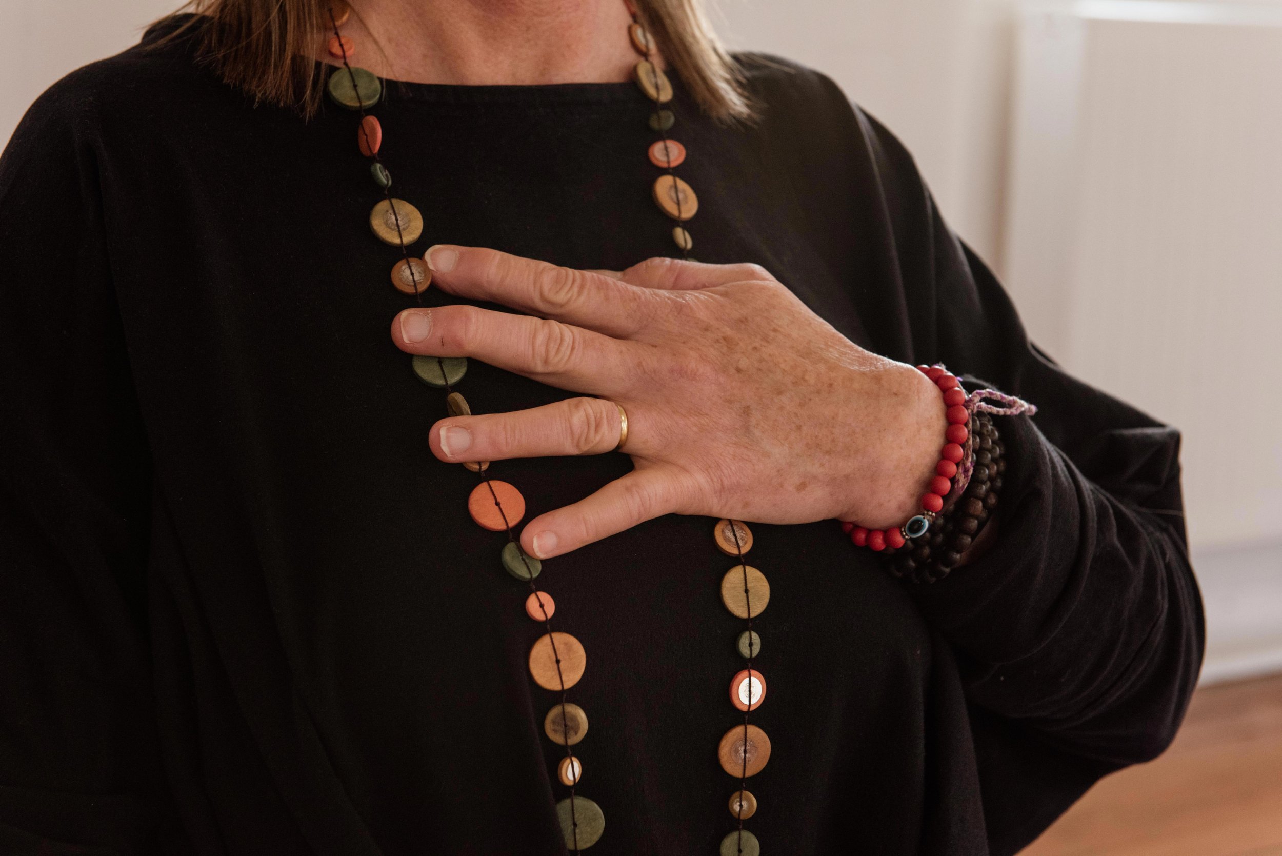 Person placing their right hand over their heart, wearing a black long-sleeve shirt, beaded necklaces, and bracelets.