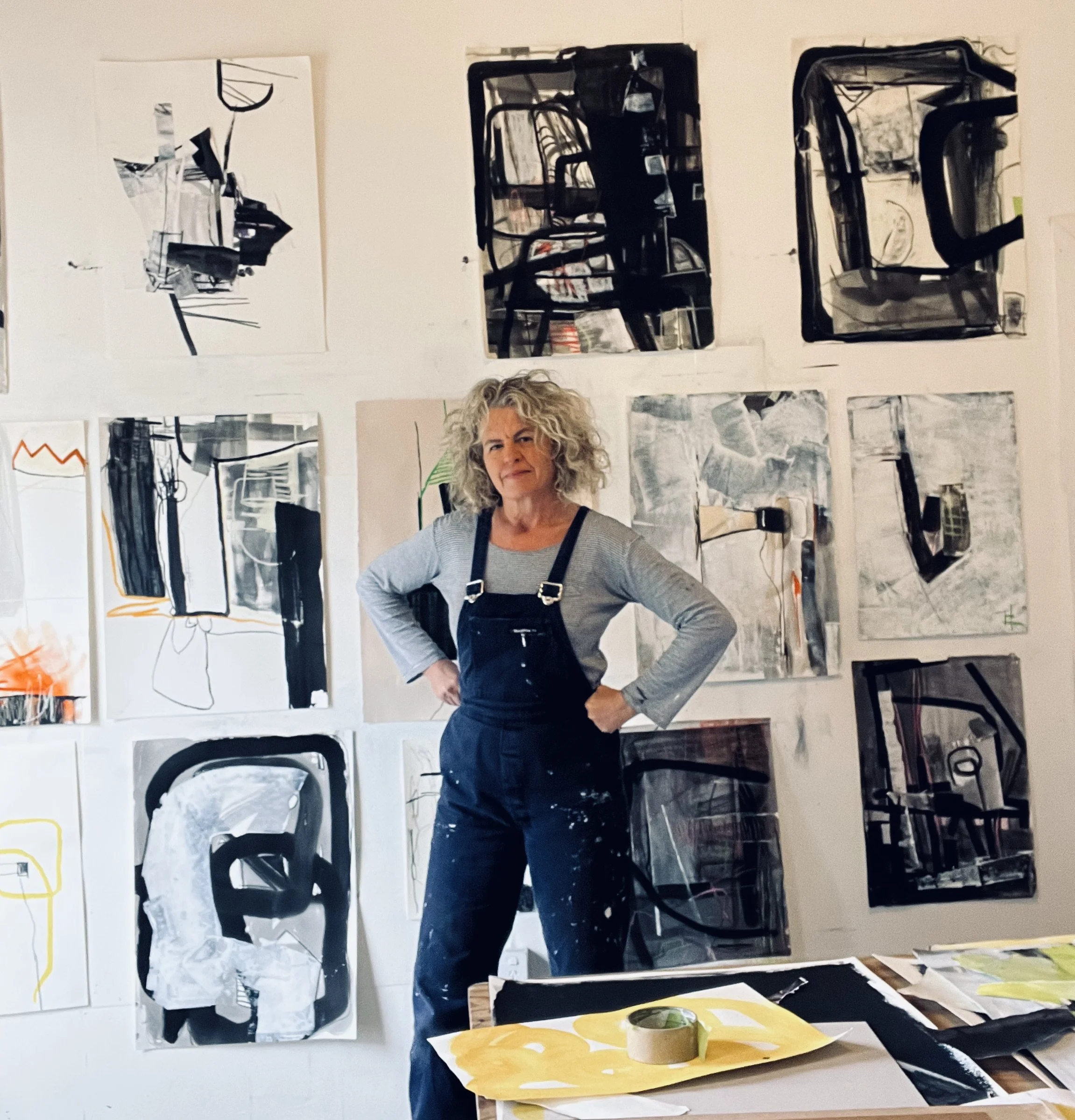 An artist standing in front of a wall filled with abstract paintings and sketches, with a table in front holding art supplies.