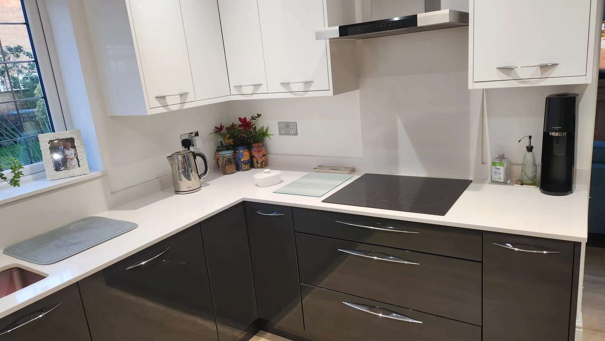 Modern kitchen corner with white cabinets, electric stovetop, kettle, countertop decor, window view, and sink.