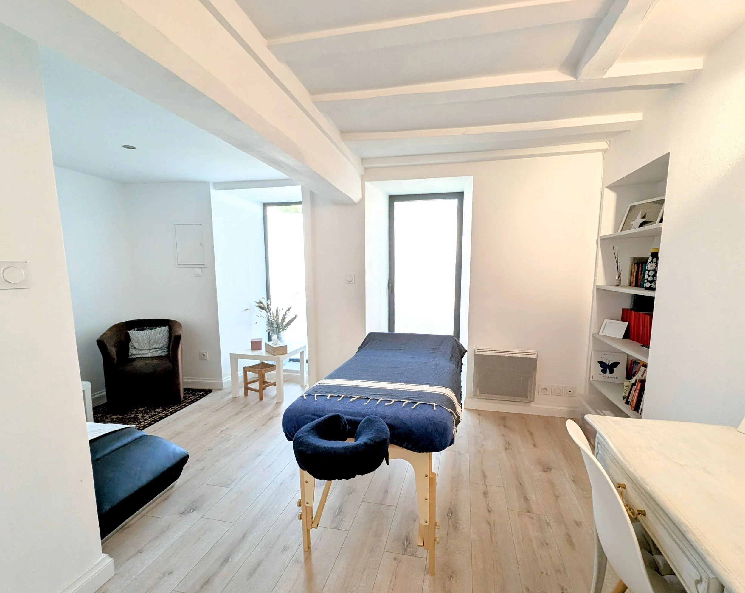 Intérieur d'un appartement avec mobilier minimaliste, comprenant un fauteuil noir, un lit, une étagère blanche avec des livres et des décorations, un tapis, et des fenêtres apportant de la lumière naturelle.