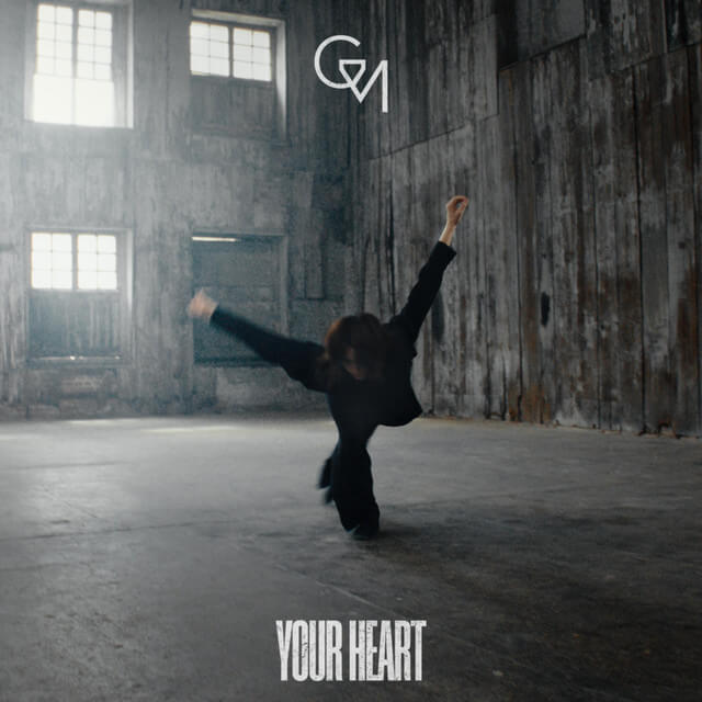 A person performing a dance move in an empty industrial warehouse with concrete floors and weathered wooden walls. The person is balancing on one hand with legs extended, wearing dark clothing. Light streams in through high windows, creating a moody