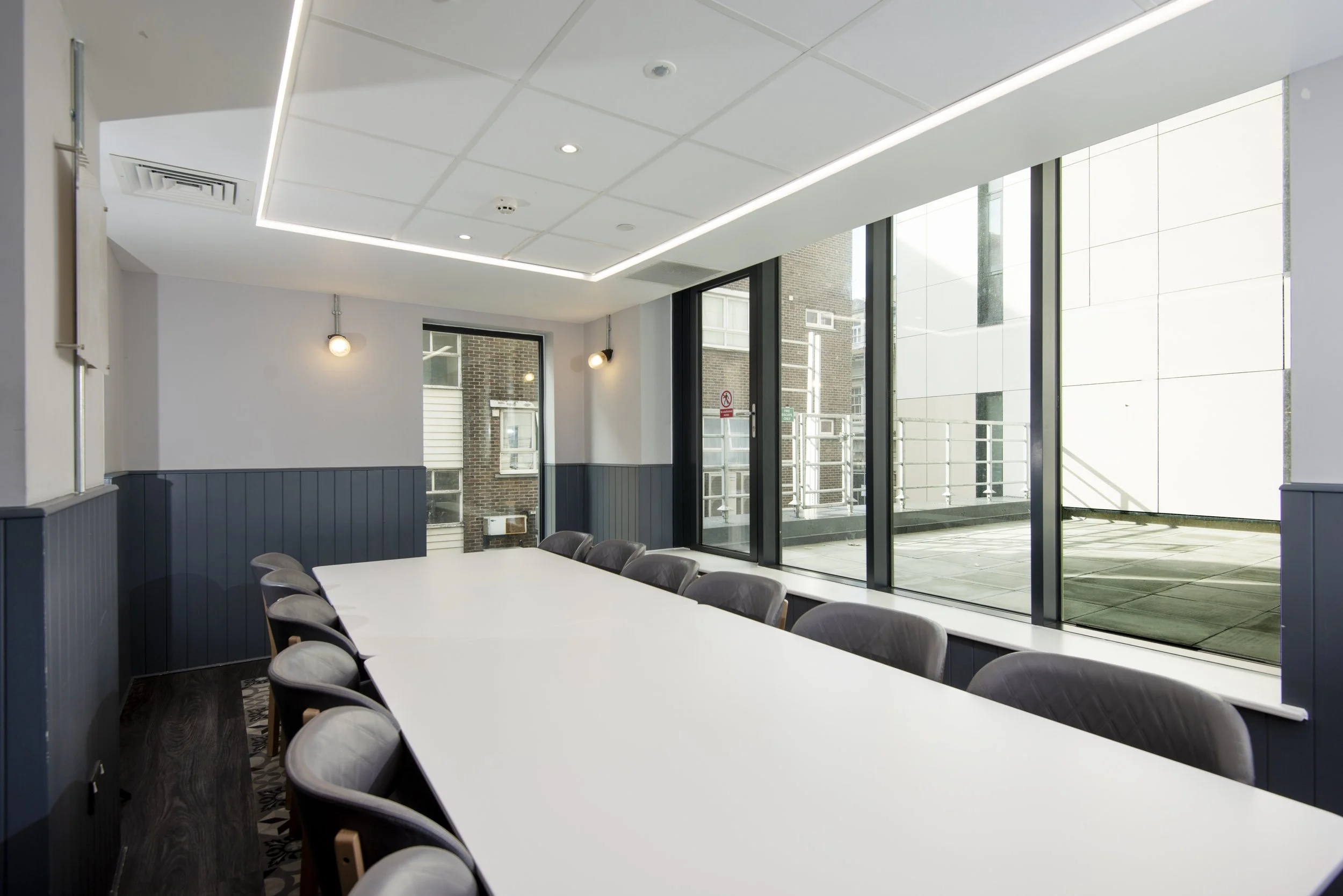 Modern conference room with a long white table, surrounded by chairs, large windows, and soft lighting.